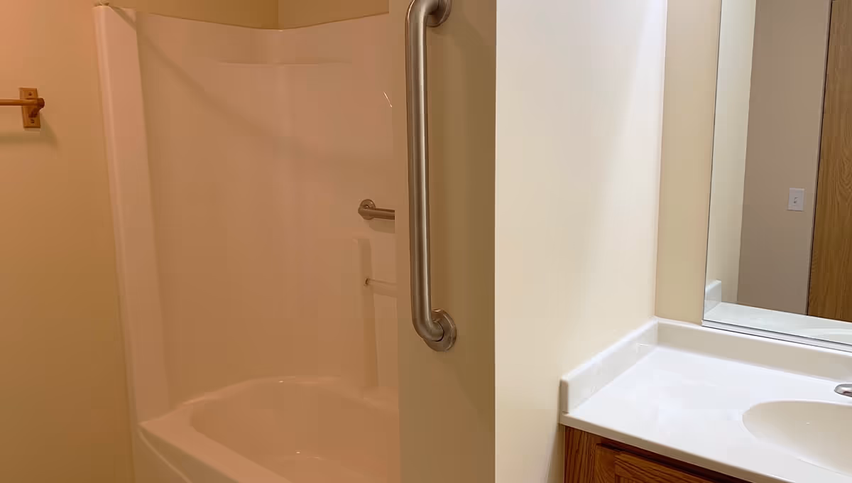 Bathroom with a white bathtub and shower combination featuring a metal grab bar. There is a white sink countertop with a faucet and a large mirror above it. The walls are painted a light beige color.