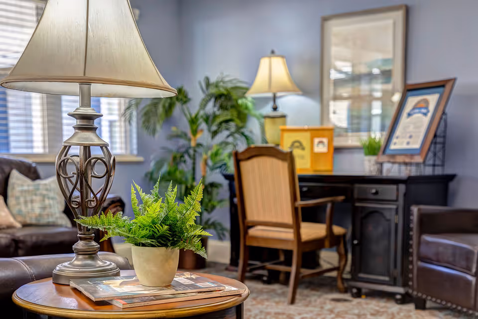 A cozy living-room lounge with a table lamp and potted fern on a side table, upholstered chairs, and a desk in the background.