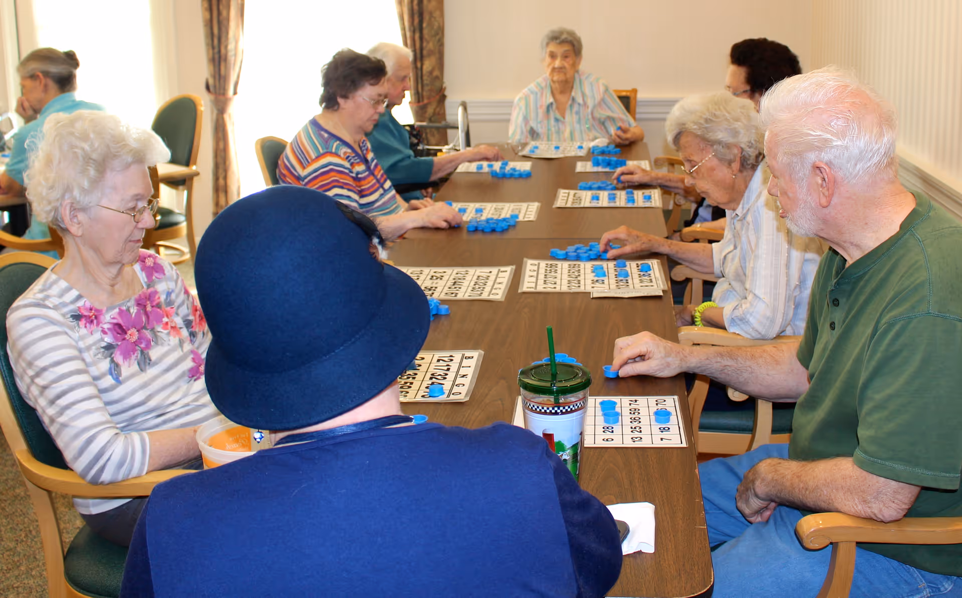 A group of elderly people sitting around a long table playing bingo. They are placing blue markers on their bingo cards. The room is well-lit with natural light coming from a window with curtains. The participants appear engaged in the game.