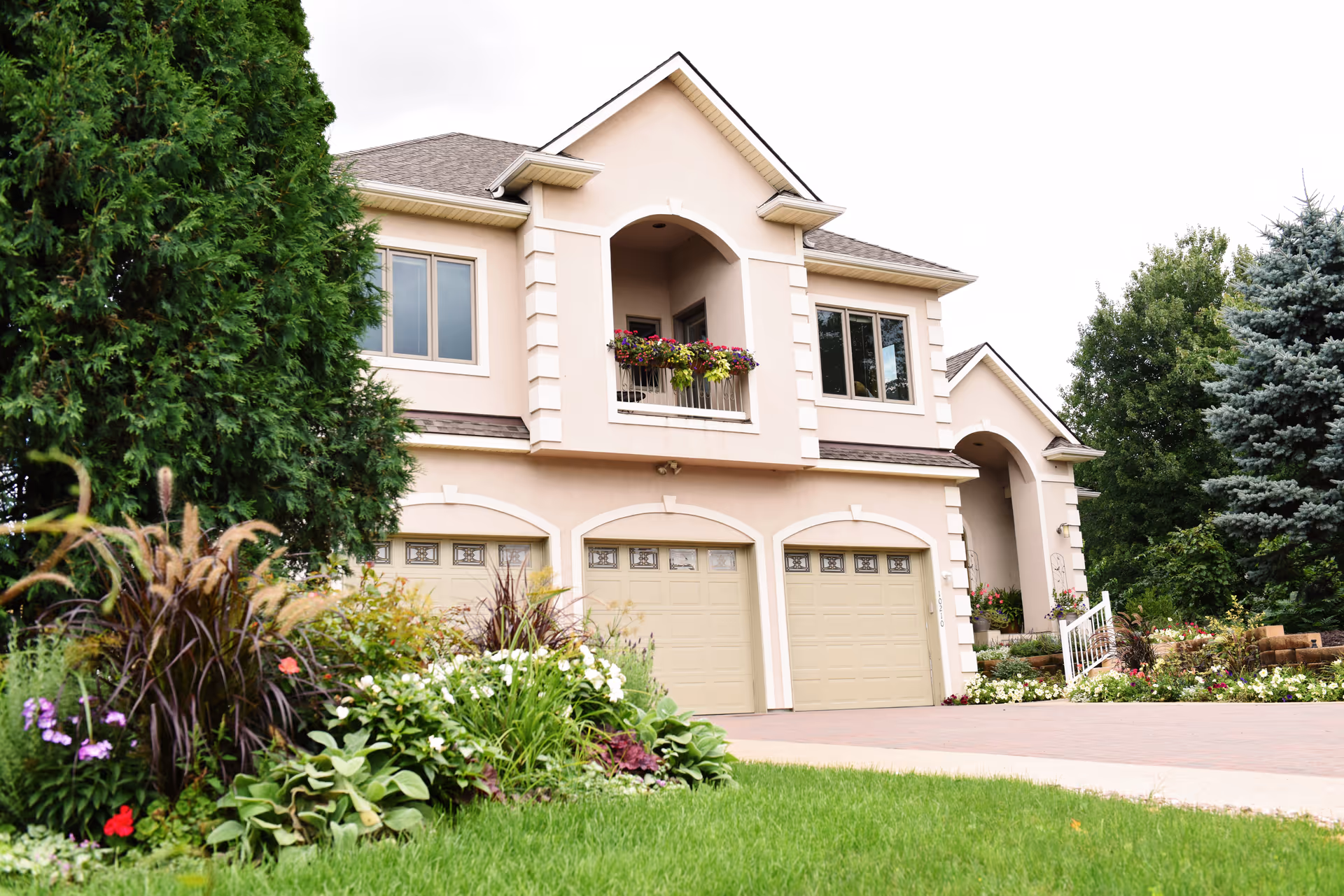 Exterior view of a two-story beige building with three garage doors, a small balcony with flower pots, and a well-maintained garden with various plants and flowers in the foreground.