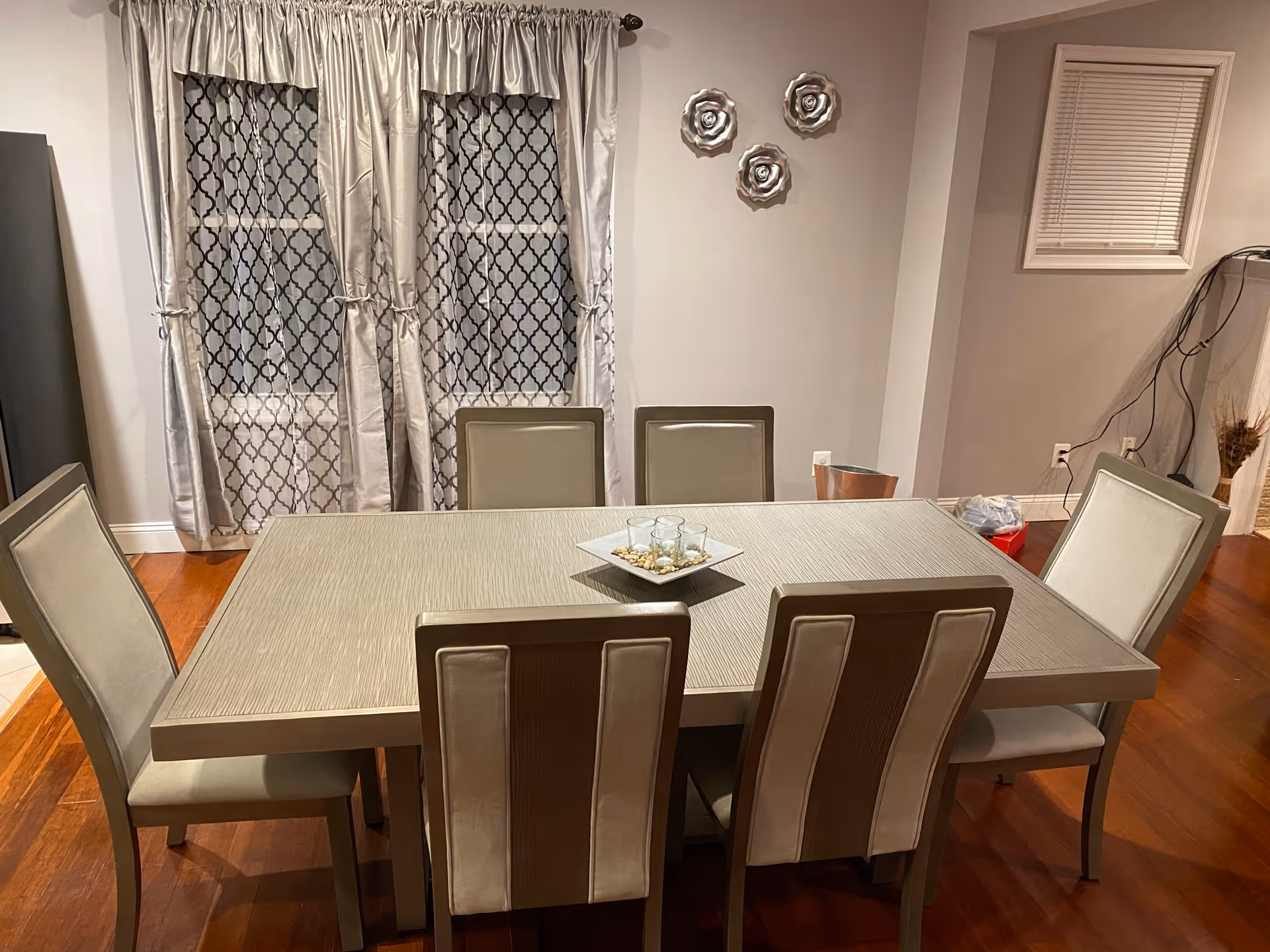 A dining room with a rectangular table surrounded by six beige upholstered chairs. The table has a decorative centerpiece with three glass candle holders on a square tray filled with small stones. Behind the table are two windows covered with patterned curtains. The room has wooden flooring and light-colored walls with three decorative wall hangings shaped like flowers.