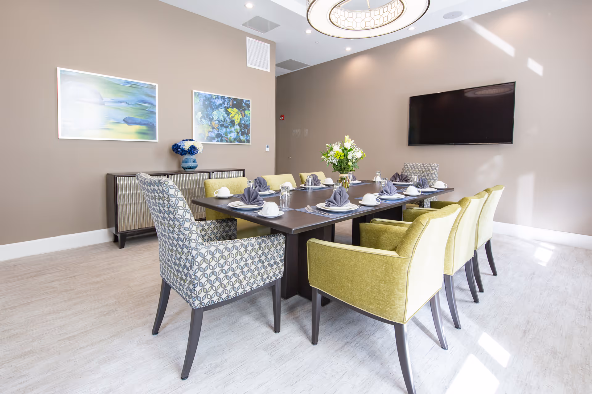 A modern dining room with a long rectangular table set for eight people. The table is arranged with white plates, cups, glasses, and gray folded napkins. There are six green upholstered chairs and two patterned armchairs at the ends. On the wall behind the table, there is a flat-screen TV mounted and two framed abstract paintings. A sideboard with a blue vase holding flowers is against the wall. The room has light-colored flooring and beige walls, with a circular ceiling light fixture above the table.