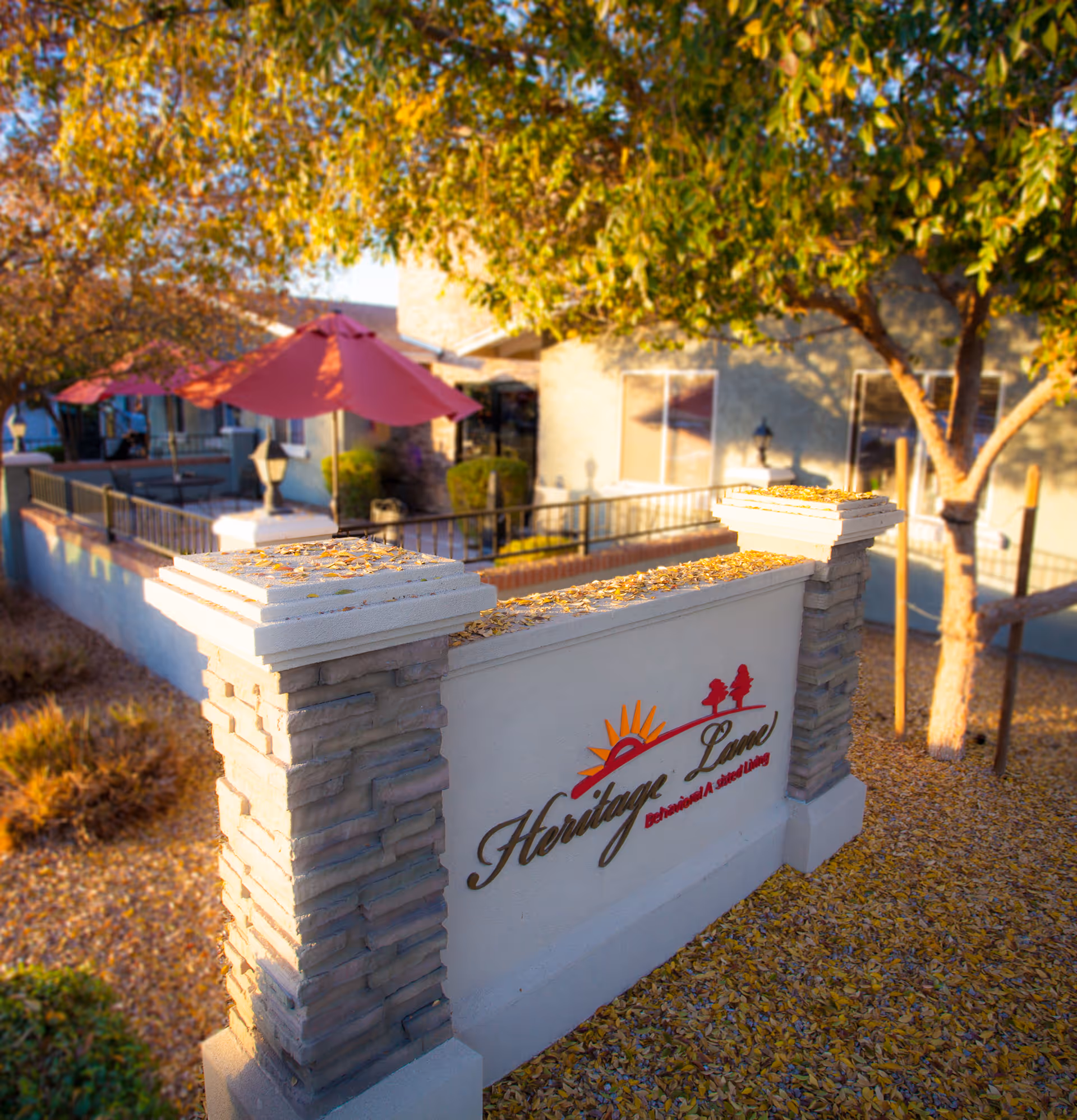 Outdoor view of Heritage Lane Behavioral Assisted Living facility sign surrounded by autumn leaves, with a patio area featuring red umbrellas and trees in the background.