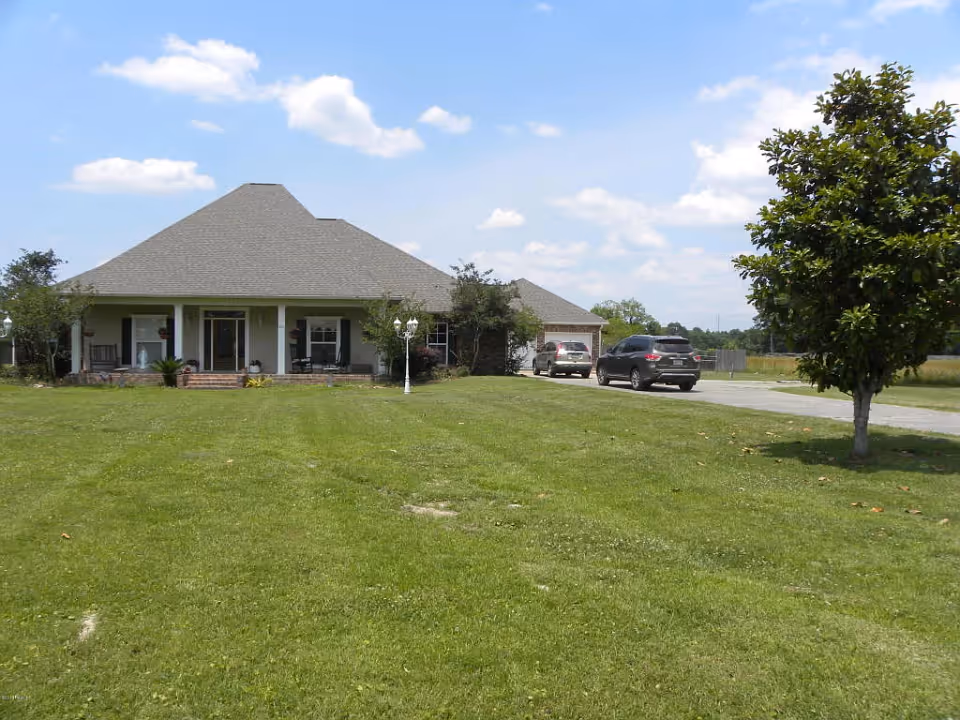 Front view of a single-story house with a wide lawn, covered porch, driveway and parked cars under a blue sky.