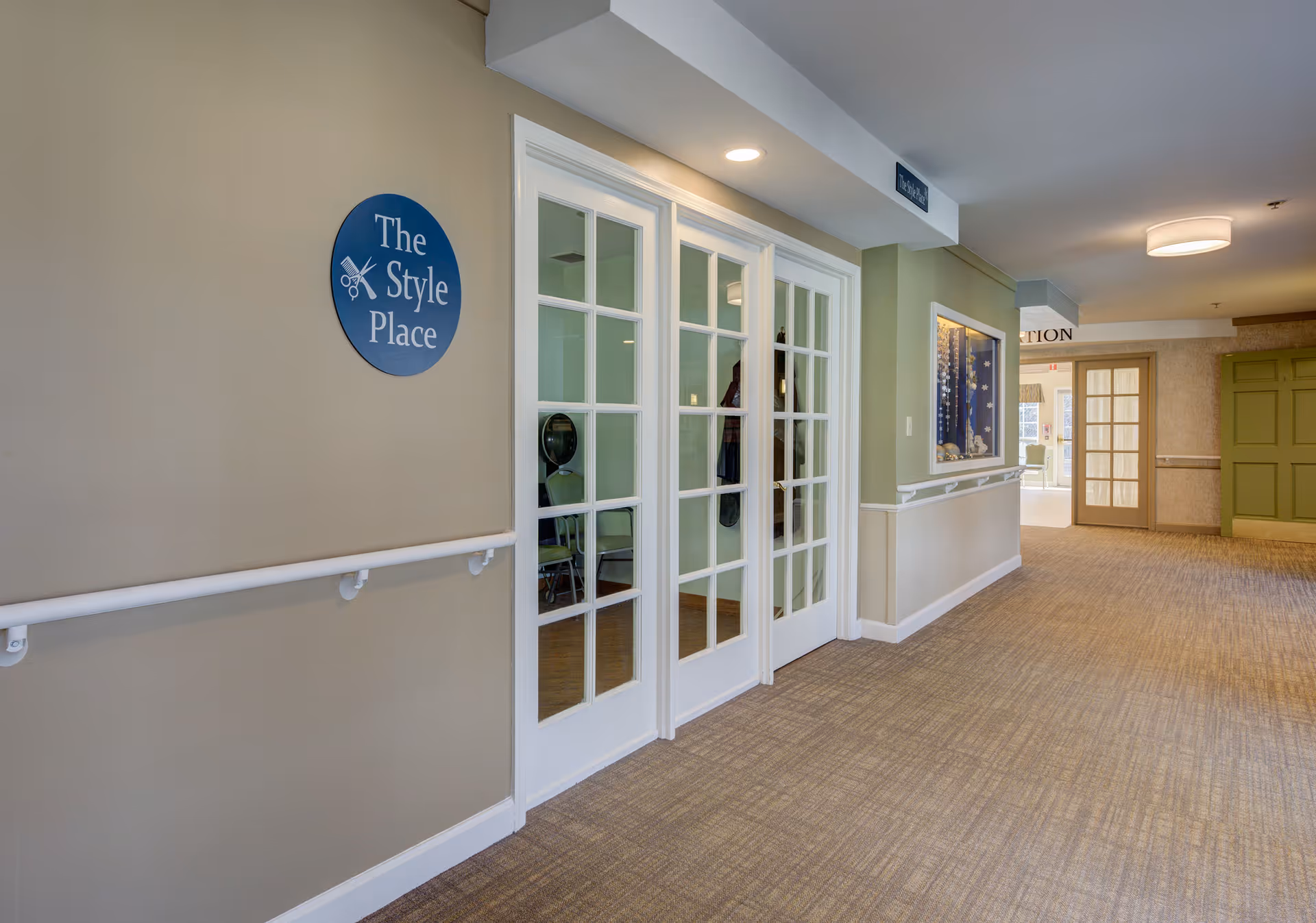 Interior hallway of a senior living facility with beige walls and carpeted floor. On the left side, there is a sign that reads 'The Style Place' next to white-framed glass double doors. The hallway is well-lit with ceiling lights and has handrails along the walls. Further down the hallway, there are additional doors and a display case mounted on the wall.