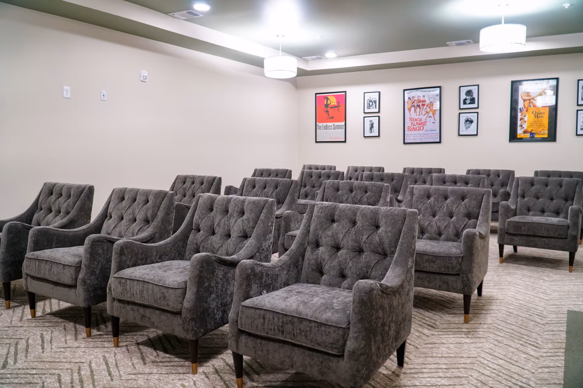 A small theater or screening room with multiple rows of gray upholstered armchairs arranged facing forward. The walls are decorated with framed movie posters and photographs. The room has a carpeted floor and ceiling lights.