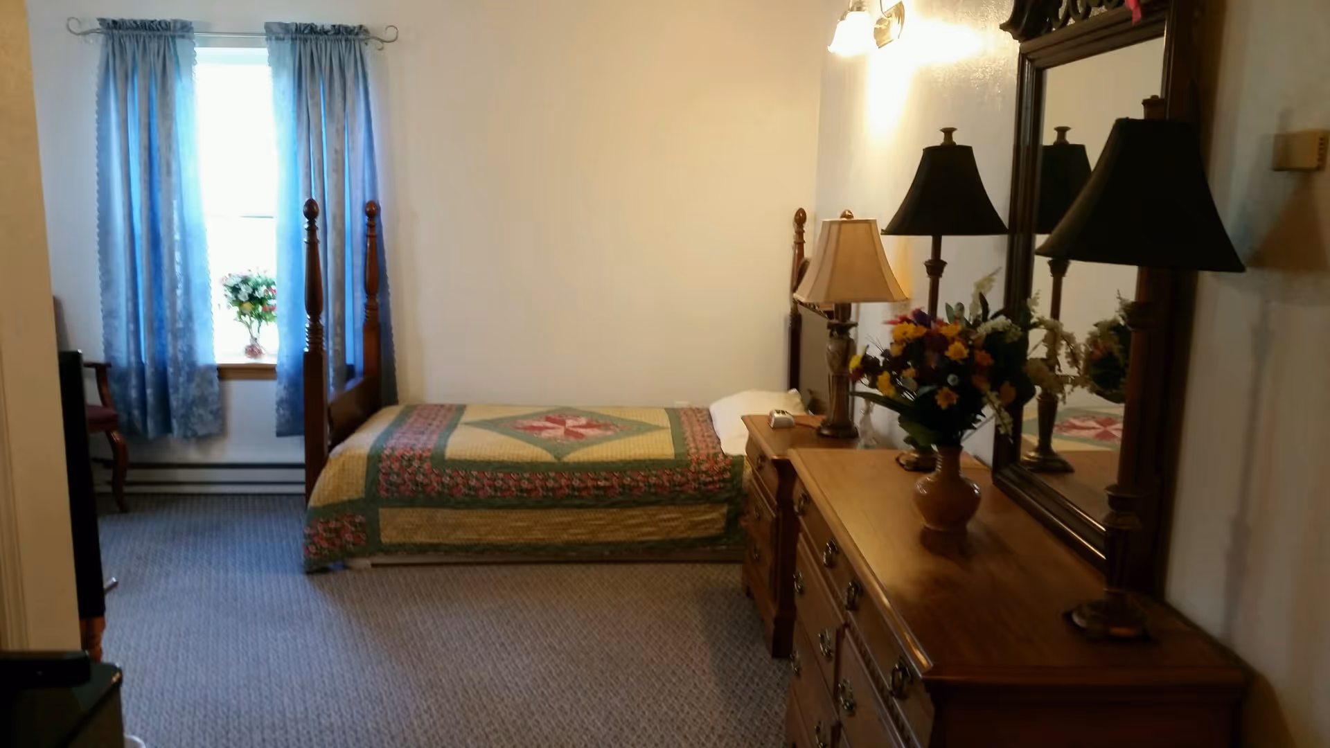 A bedroom with a single wooden bed covered with a patterned quilt, a wooden dresser with a large mirror, two table lamps, and a vase of colorful flowers. Blue curtains hang on a window letting in natural light.