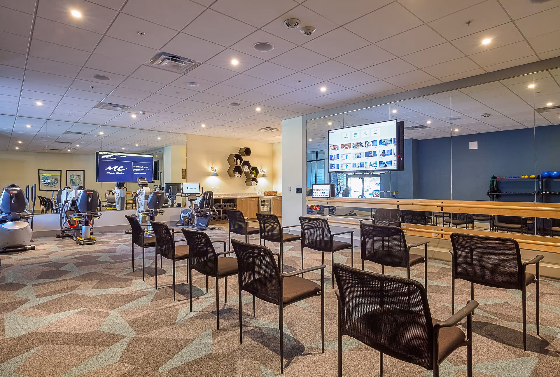 Spacious fitness/activity room with rows of chairs, exercise machines, mirrored wall, and wall-mounted TVs.
