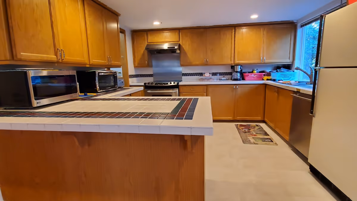 A kitchen with wooden cabinets, a tiled countertop island, a stainless steel stove with a range hood, a microwave, a toaster oven, a coffee maker, a refrigerator, a dishwasher, and a window letting in natural light.