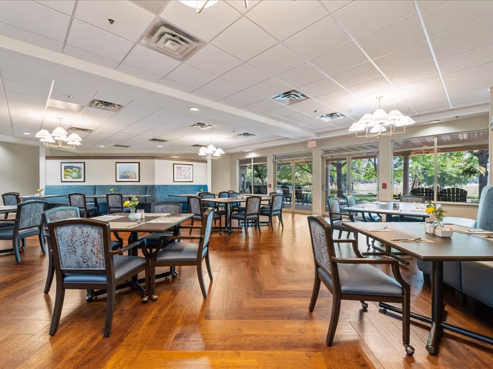 Spacious dining room with multiple tables and chairs and large windows looking out to trees and outdoor seating.