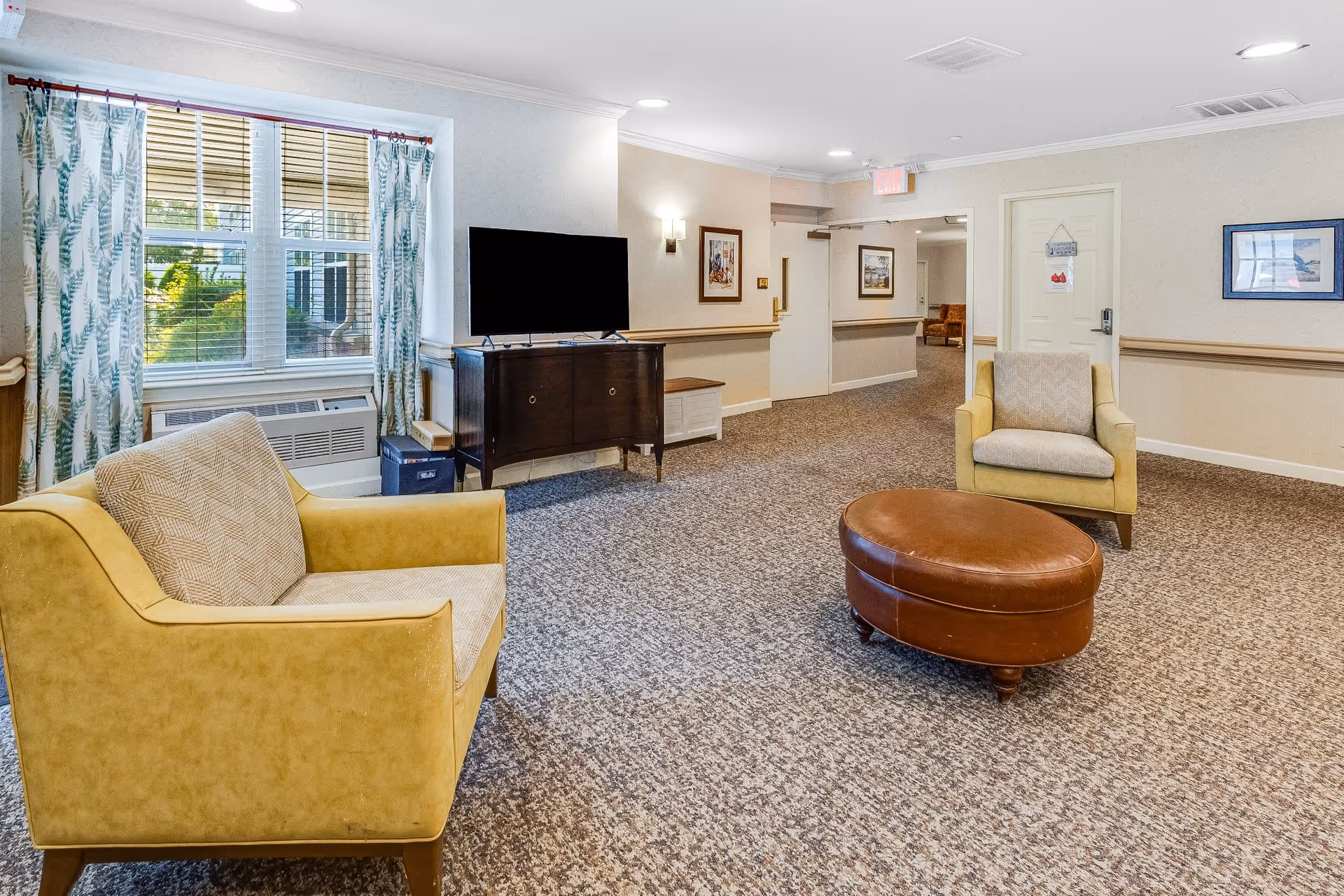 A cozy common area in a senior living facility with two yellow armchairs, a round brown leather ottoman, a flat-screen TV on a dark wooden cabinet, patterned curtains on a window, and framed artwork on beige walls.