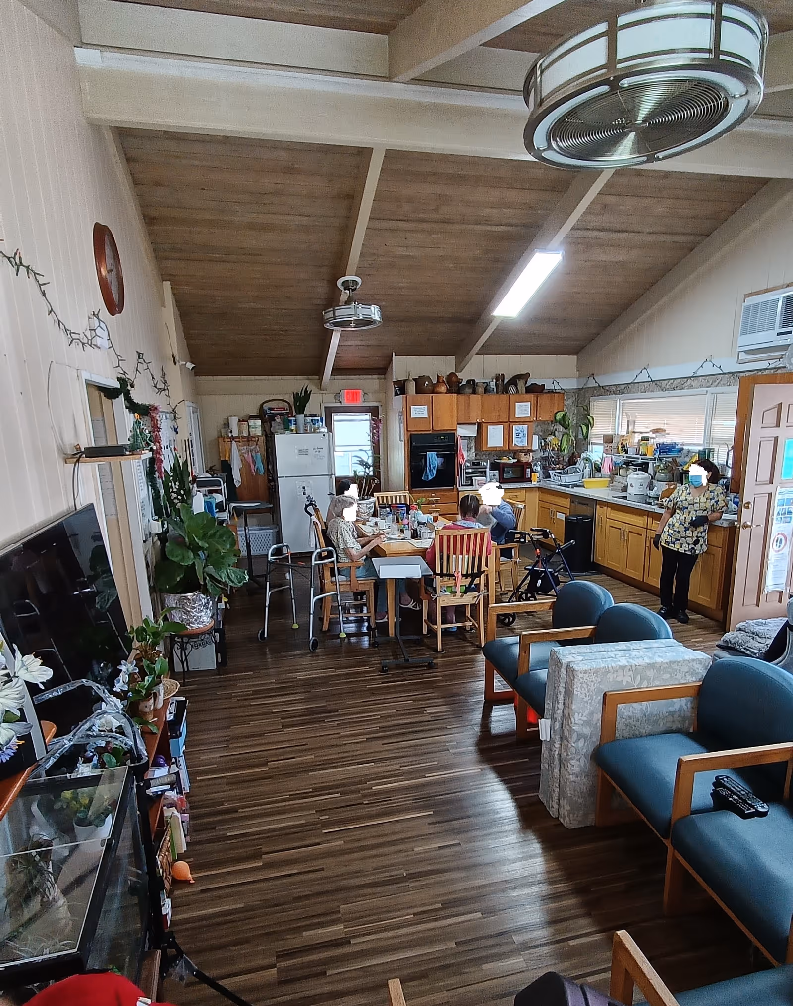Interior view of a senior living facility common area with a kitchen and dining space. Several elderly individuals are seated around a table engaged in activities, with a caregiver standing nearby. The room has wooden flooring, a high wooden ceiling with beams, ceiling fans, and various plants and decorations. There are chairs and a television on one side, and kitchen cabinets, a refrigerator, and appliances on the other.