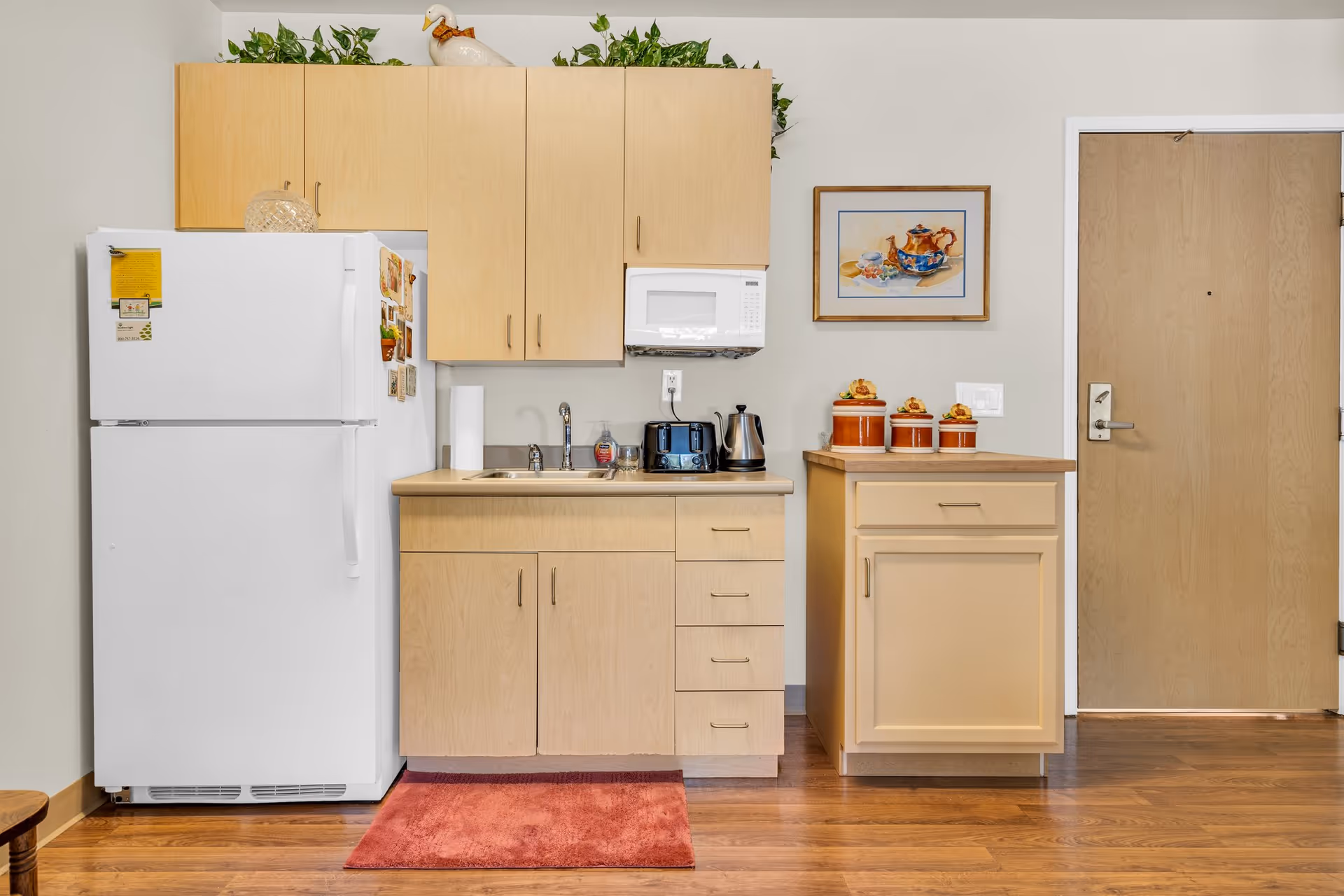 A small kitchen area with light wood cabinets, a white refrigerator with magnets, a microwave mounted above the counter, a stainless steel sink, a toaster, an electric kettle, and three decorative canisters on a separate cabinet. A framed painting hangs on the wall next to a wooden door. The floor is wood with a red rug in front of the sink.