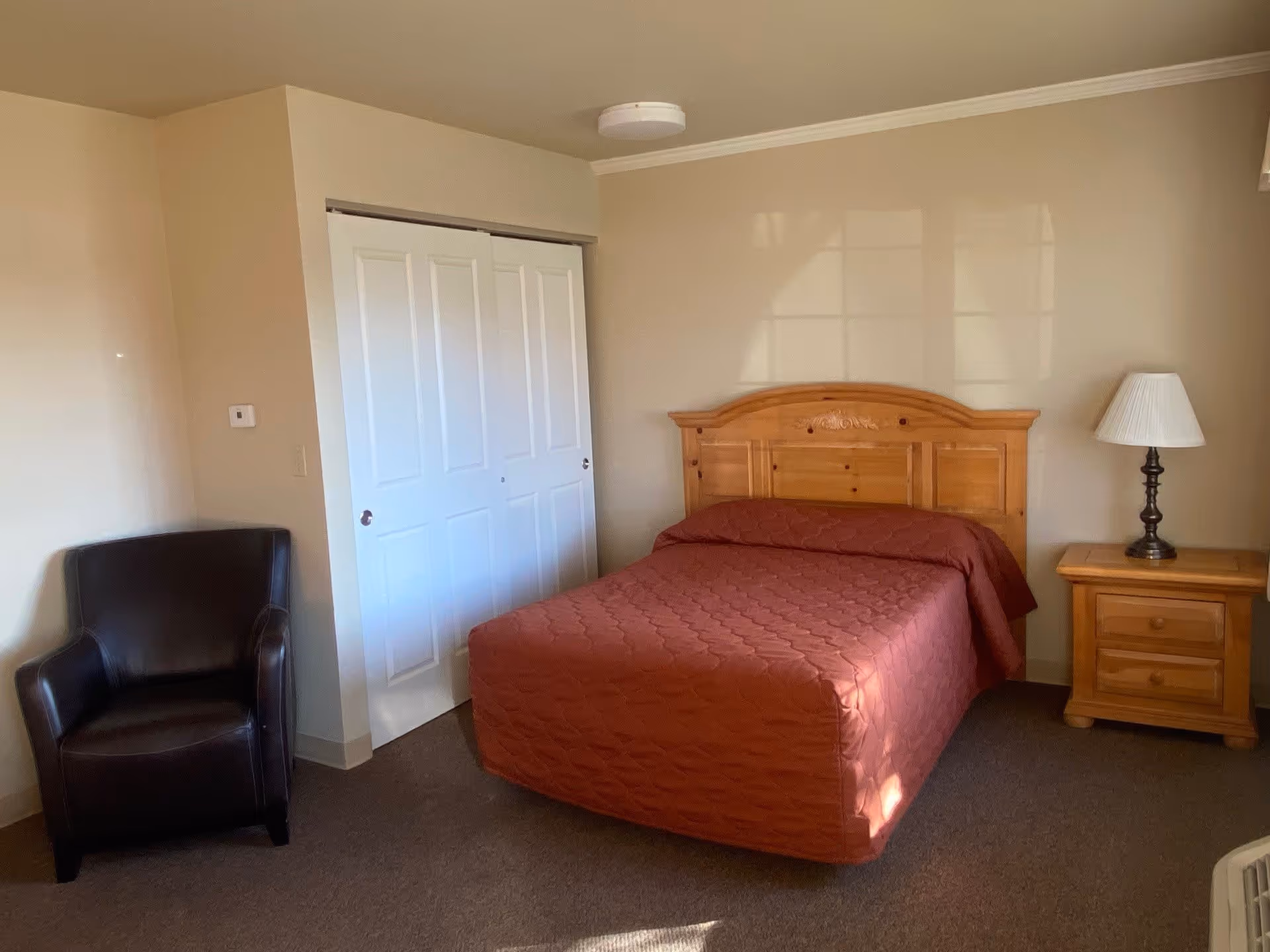 A bedroom with a wooden bed frame and a red quilted bedspread. There is a wooden nightstand with a lamp on the right side of the bed and a dark brown armchair in the corner near a white closet with sliding doors. The walls are beige and the floor is carpeted.