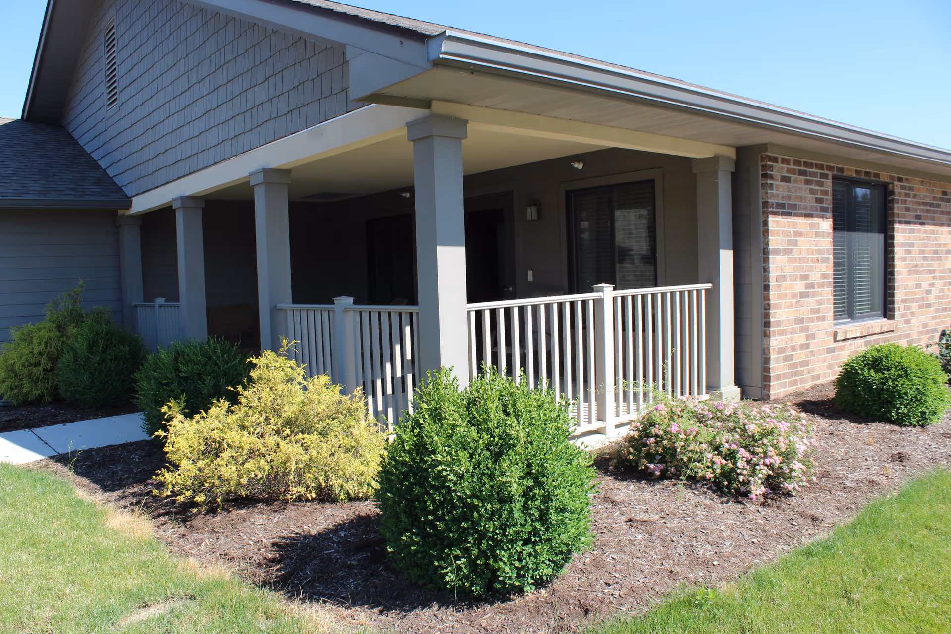 Exterior view of a single-story building with a covered porch supported by columns. The porch has a white railing and is surrounded by neatly trimmed bushes and landscaping. The building features a combination of brick and siding with windows visible.