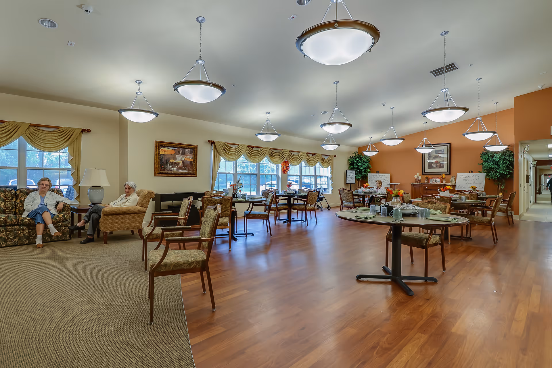 A spacious assisted living common area with wooden floors and multiple round tables set with cups and plates. Two elderly women are seated on floral and beige armchairs near a window with yellow curtains. The room is well-lit with multiple hanging ceiling lights and decorated with framed paintings and potted plants.