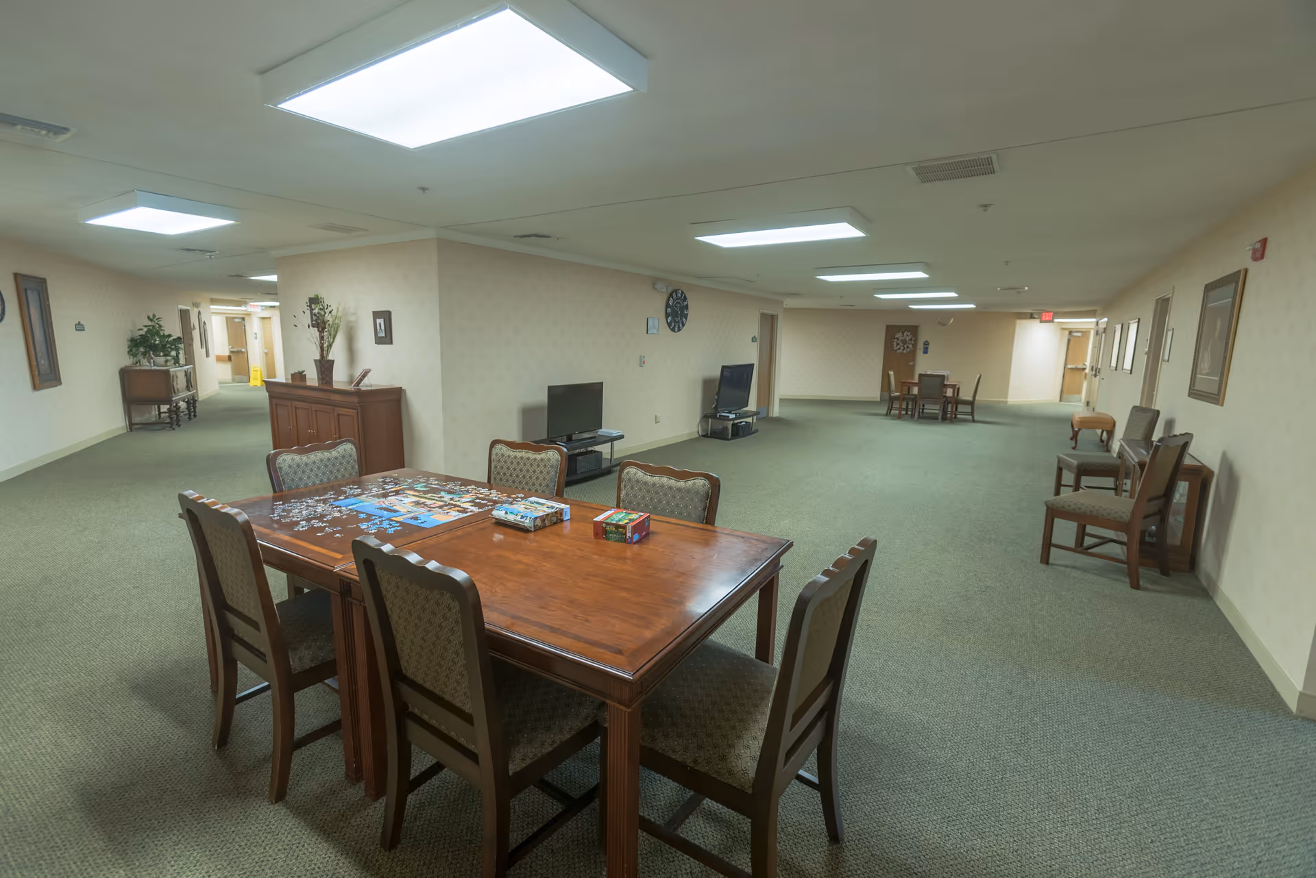 A spacious common area in a senior living facility with a wooden table and six chairs in the foreground. On the table, there is a partially completed jigsaw puzzle and two puzzle boxes. The room has green carpet, beige wallpaper, and several chairs lined up along the walls. Two televisions are placed against the walls, and there are framed pictures and a clock on the walls. The area is well-lit with ceiling lights and has multiple doorways leading to other rooms.