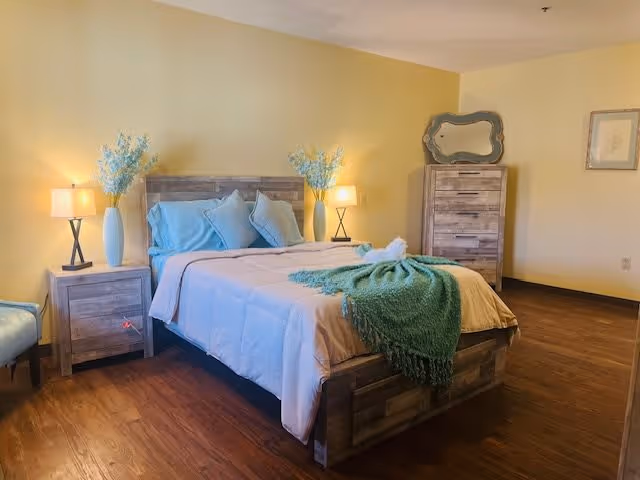 Well-lit bedroom with a wooden bed, matching nightstands and dresser, bedside lamps, and a green throw on the bed against pale yellow walls.