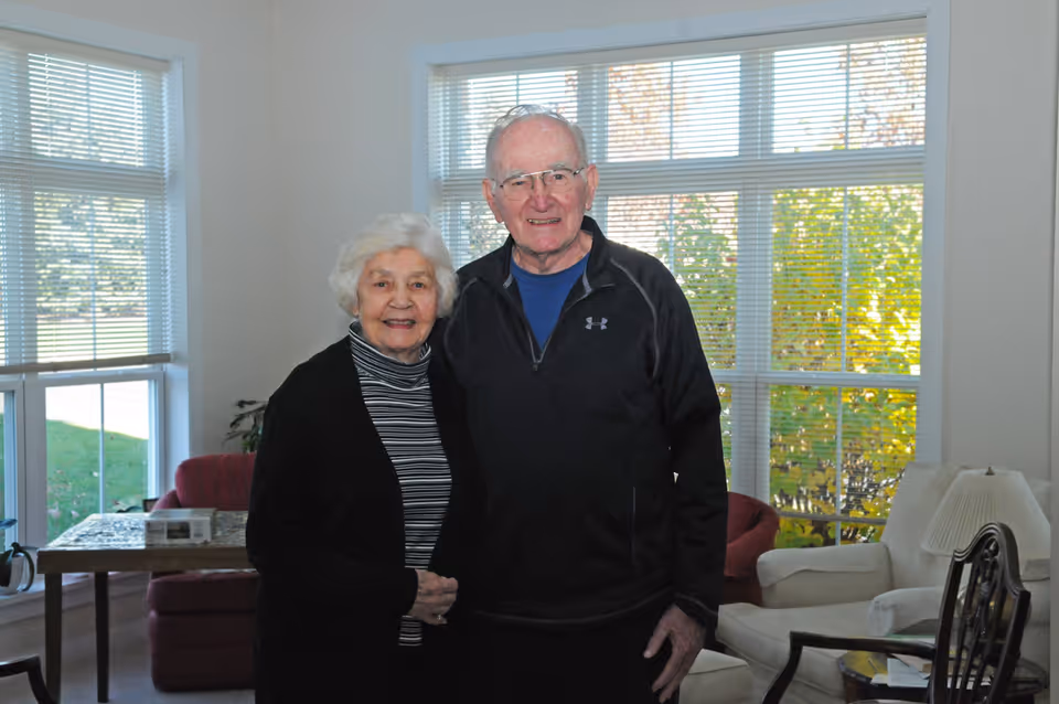 Two smiling older adults standing together in a bright living room with large windows, armchairs, and a side table.
