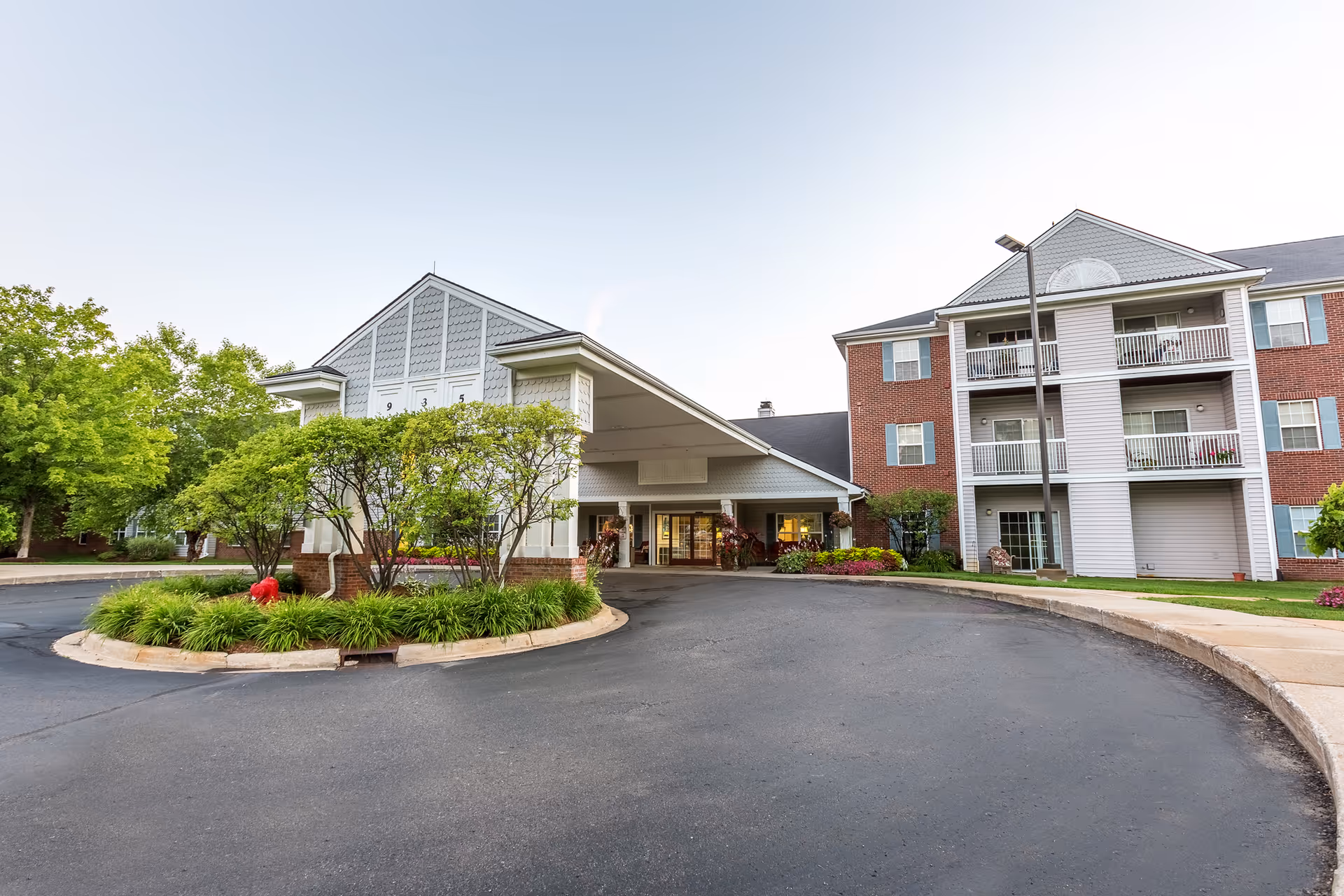 Exterior view of Independence Village of White Lake showing a multi-story residential building with balconies and a covered entrance driveway surrounded by landscaped greenery and trees under a clear sky.