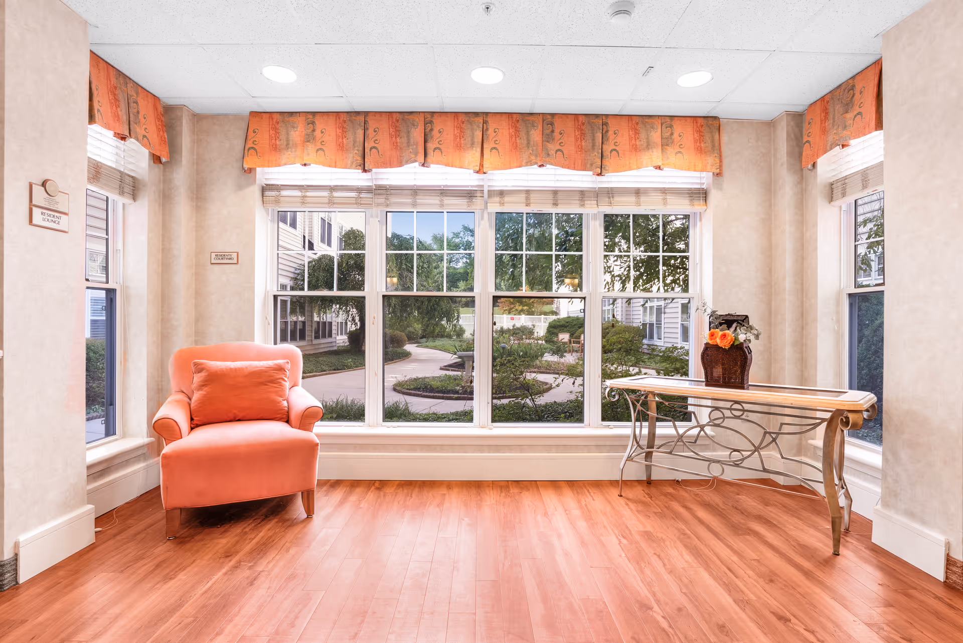 A bright room with large windows overlooking a garden path outside. Inside, there is a coral-colored armchair with a matching pillow on the left and a decorative glass and metal table with a flower arrangement on the right. The room has light-colored walls, wood flooring, and orange valances above the windows.