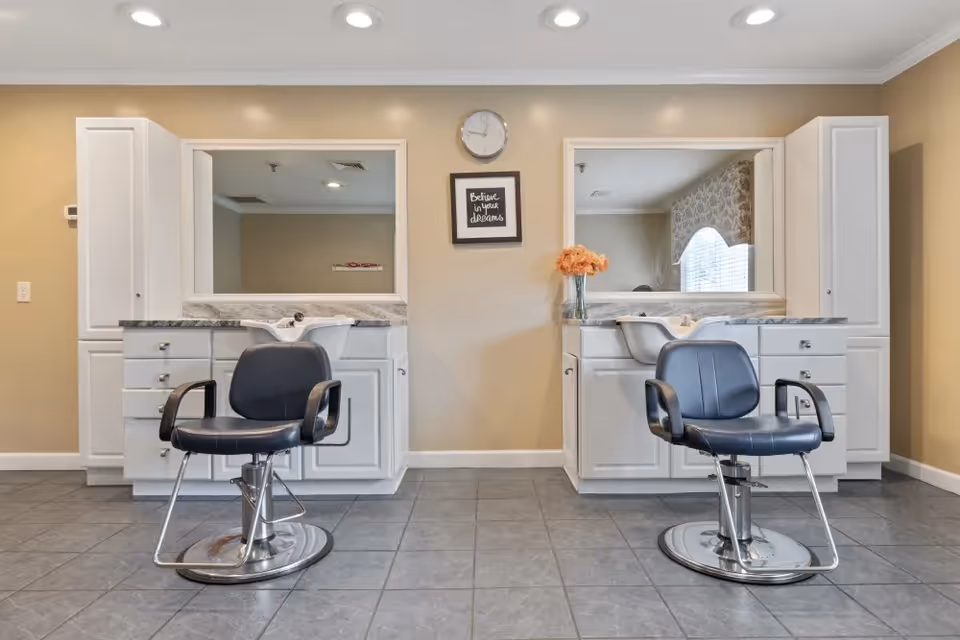 Interior view of a salon area with two black salon chairs in front of white cabinets and sinks. Each station has a large mirror above the sink. A clock and a framed sign that reads 'Believe in your dreams' are mounted on the beige wall between the two stations. A vase with orange flowers is placed on the right counter.