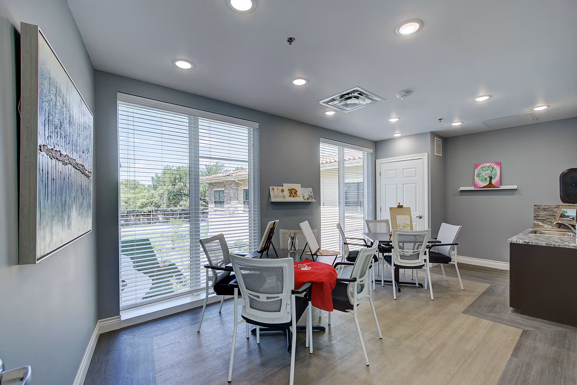 A bright and modern common area with large windows covered by white blinds letting in natural light. The room has several white chairs around small tables, some with art supplies and easels, suggesting an activity or craft space. The walls are painted gray and decorated with colorful artwork. The floor has a mix of light and dark wood patterns, and there is a small kitchenette area with a countertop and sink.