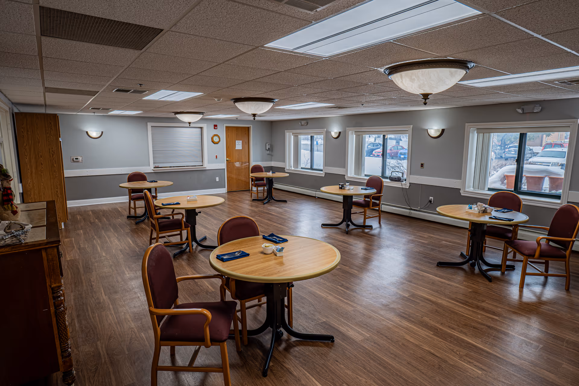 A dining room with five round wooden tables, each accompanied by two to three maroon cushioned chairs. The room has wood flooring, gray walls with white trim, several windows showing a parking lot outside, and ceiling lights providing illumination.