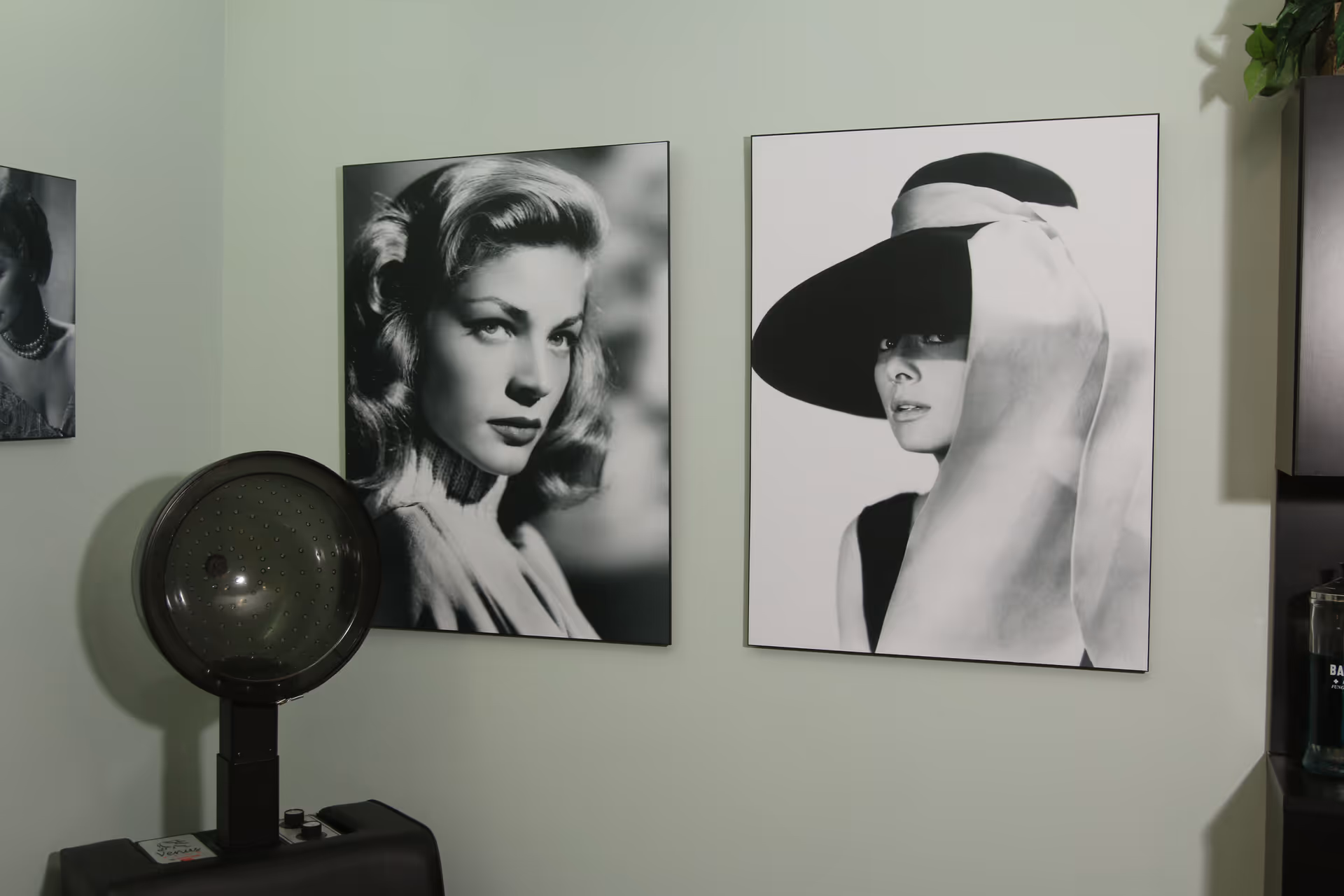 A corner of a room with two large black and white framed portraits of women on a light green wall. One woman has styled hair and is looking to the side, while the other wears a large hat with a ribbon and looks forward. Below the portraits is a vintage hair dryer machine. Part of a dark cabinet and a bottle are visible on the right side.