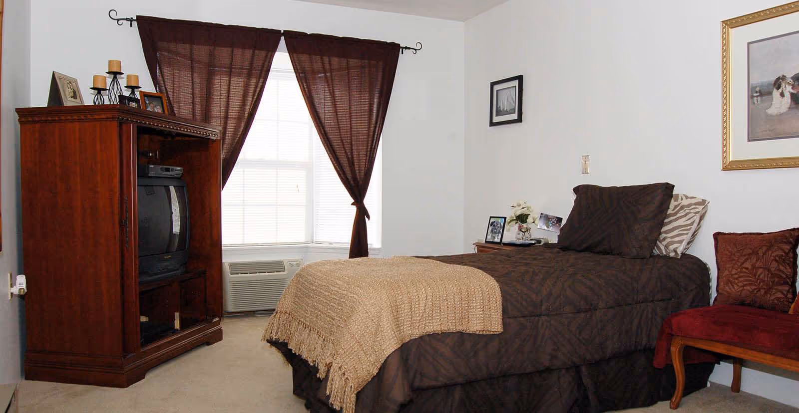 Cozy single bedroom with a twin bed in brown bedding, a wooden entertainment cabinet with TV, a bay window with brown curtains, and a small upholstered chair.