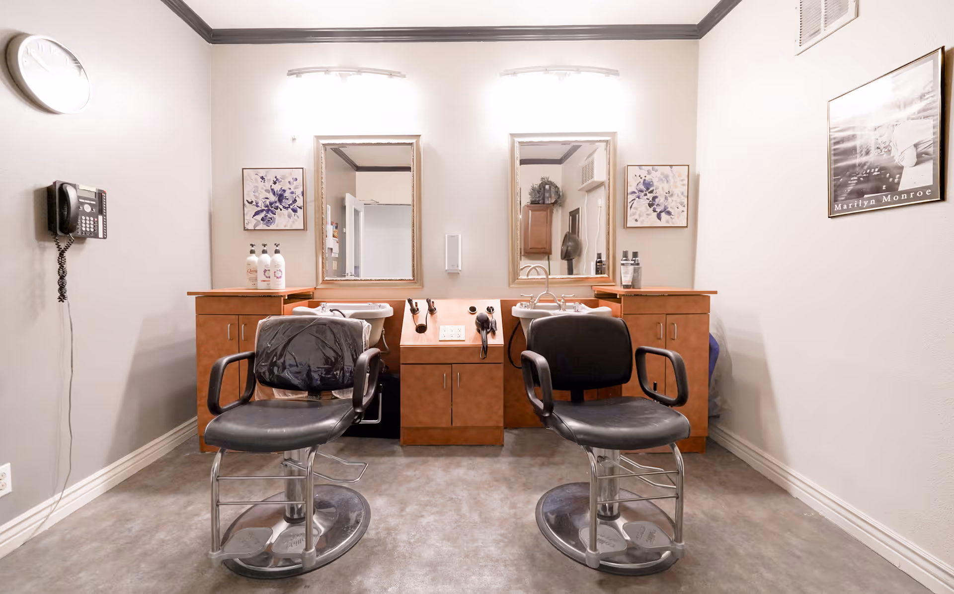 A small salon area in an assisted living facility with two black salon chairs in front of two mirrors mounted on a wooden counter. The counter has hair care products, a hair dryer, and a sink. The walls are light-colored with a clock, a telephone, and framed pictures including one of Marilyn Monroe.