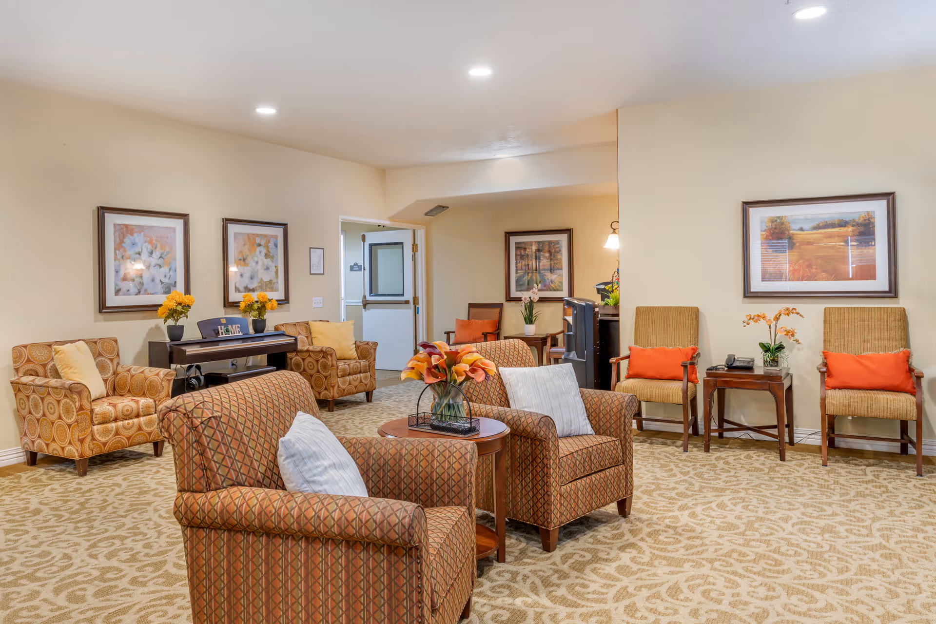 A well-lit senior living common room with upholstered chairs, side tables, floral arrangements, and framed artwork on the walls.