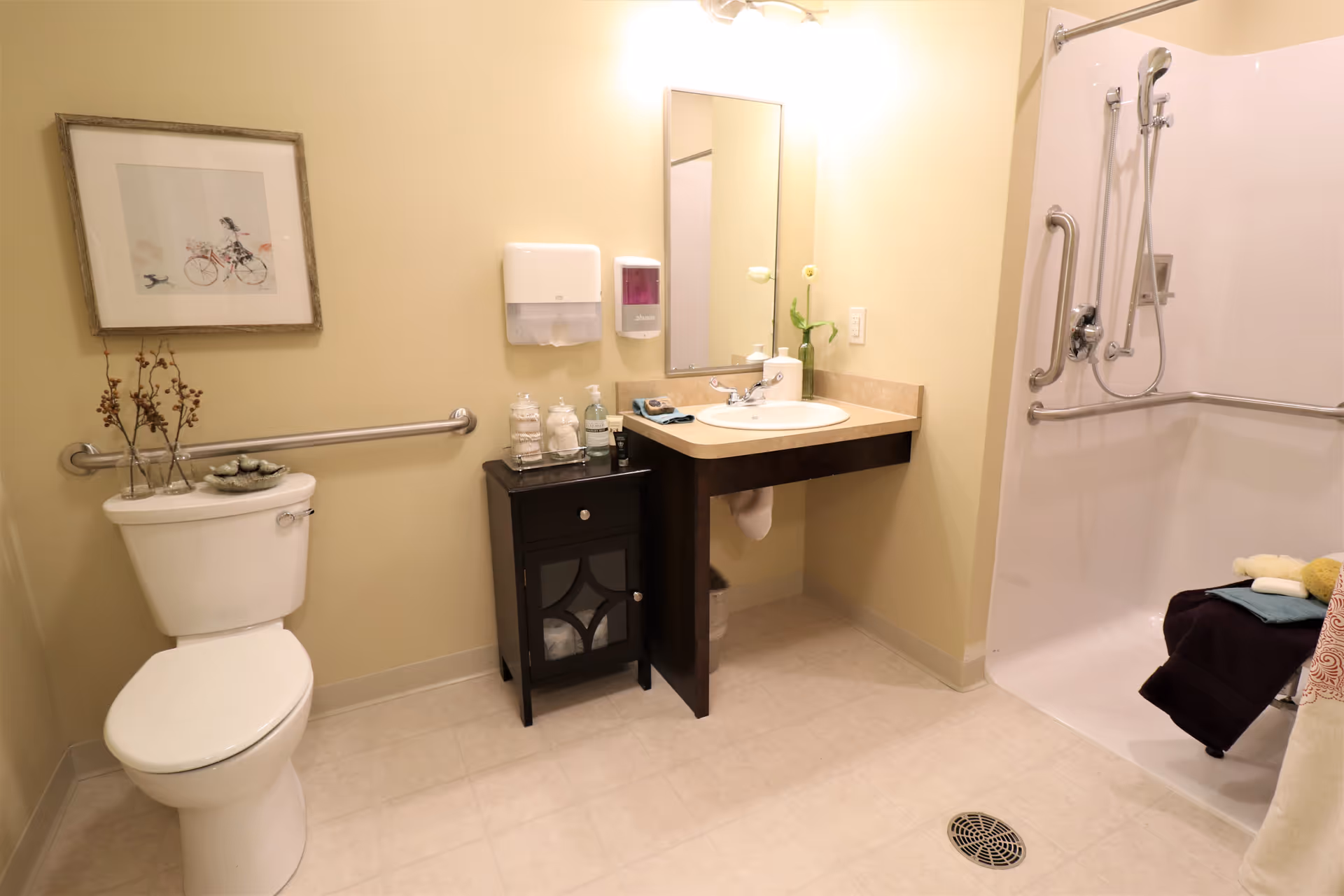 A clean and accessible bathroom with a white toilet, a small black cabinet with toiletries, a wall-mounted sink with a mirror above it, and a walk-in shower with grab bars and a handheld showerhead. The walls are painted light yellow and there is a framed picture above the toilet.