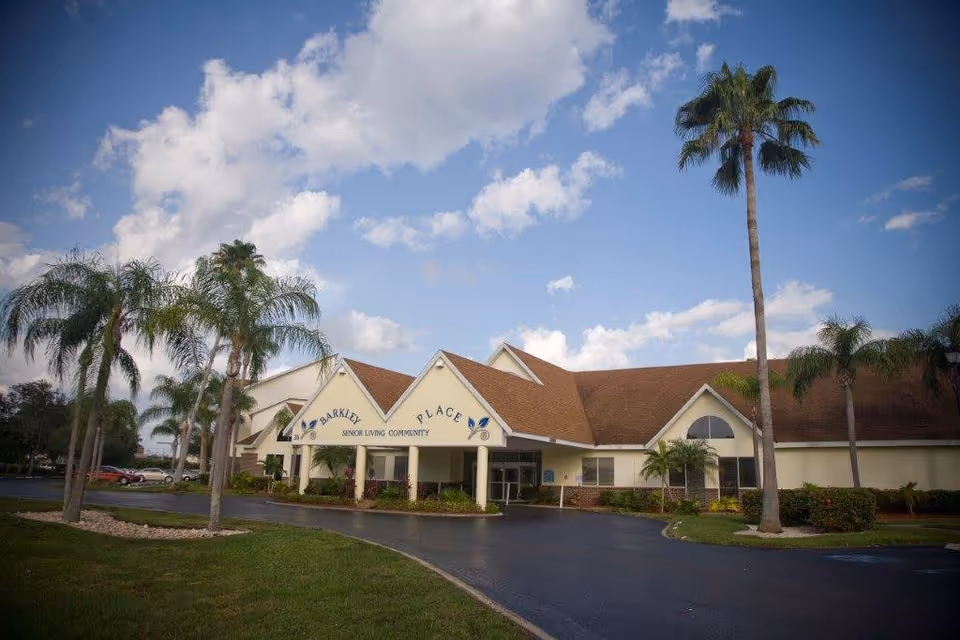Exterior view of Merrill Gardens at Barkley Place senior living community building with a driveway, palm trees, and a partly cloudy sky.