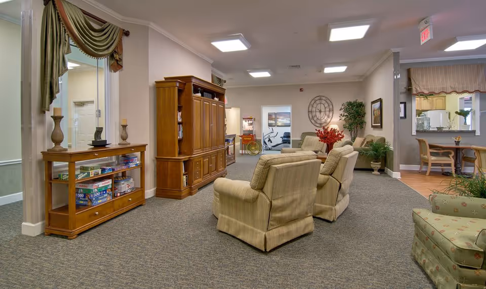 A cozy senior living common area with beige armchairs arranged around a wooden entertainment center. There is a wooden console table with board games underneath, decorative candles, and a large mirror above it. The room has carpeted flooring, neutral-colored walls, and ceiling lights. In the background, there is a small dining area with a table and chairs, a large wall clock, plants, and framed artwork.