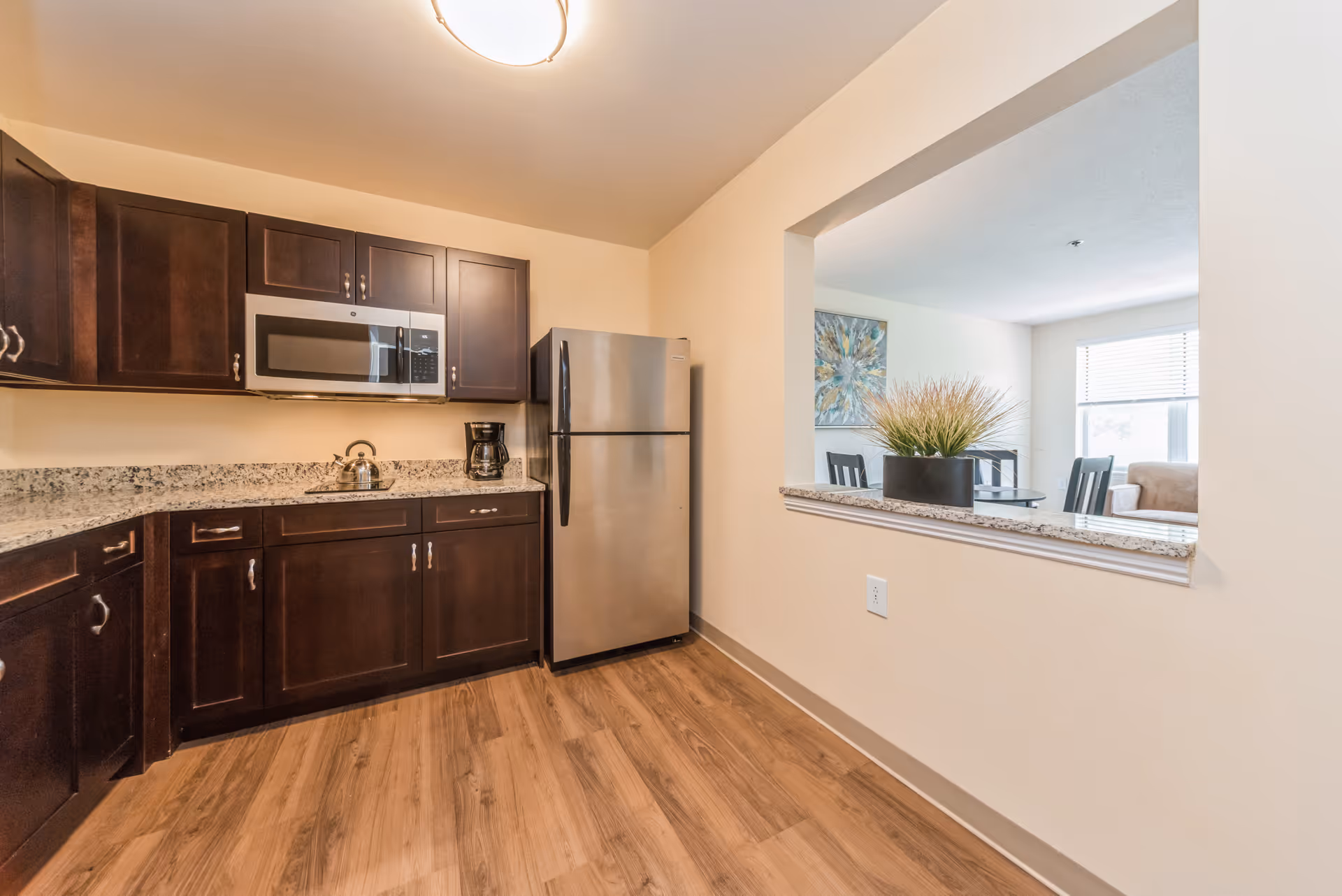A modern kitchen with dark wood cabinets, granite countertops, a stainless steel refrigerator, microwave, coffee maker, and a kettle. The kitchen has wood flooring and a pass-through window opening to a dining and living area with a plant on the counter ledge.