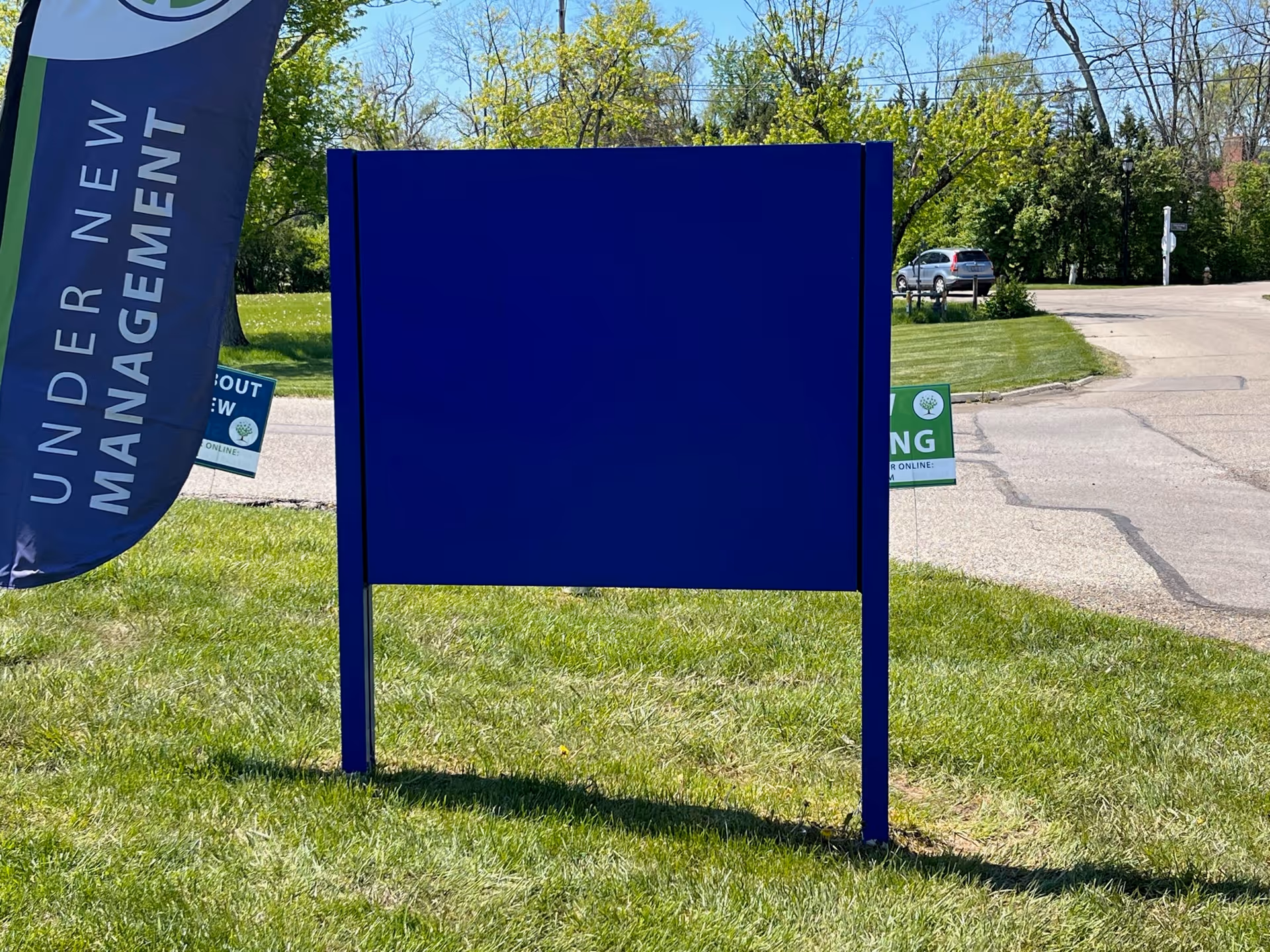 A blank blue signboard standing on a grassy area near a road. To the left of the signboard is a vertical flag with the text 'UNDER NEW MANAGEMENT'. In the background, there are trees, a parked car, and a clear blue sky.