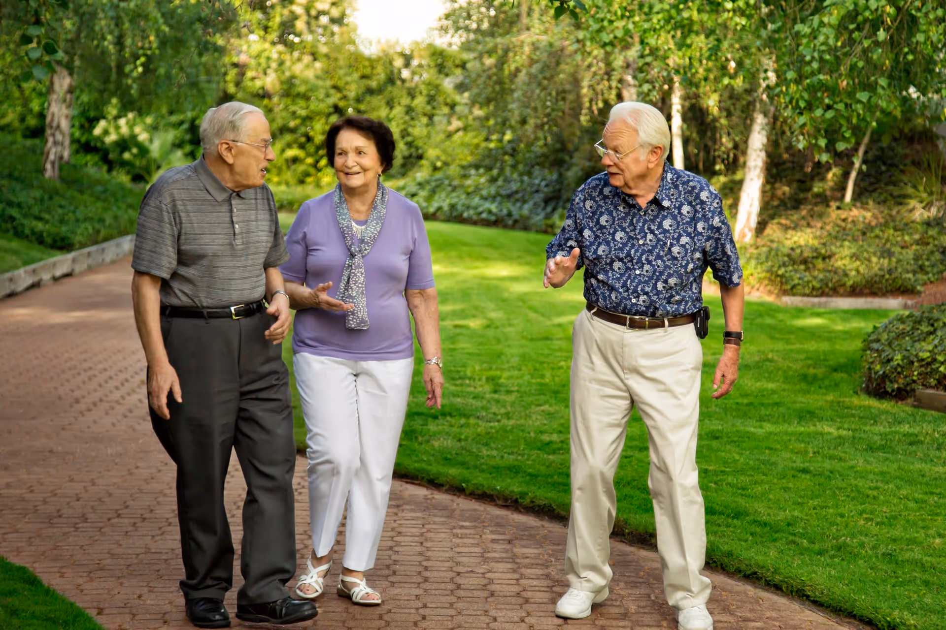 Three elderly people walking and talking on a paved path in a lush green garden area with trees and bushes in the background.