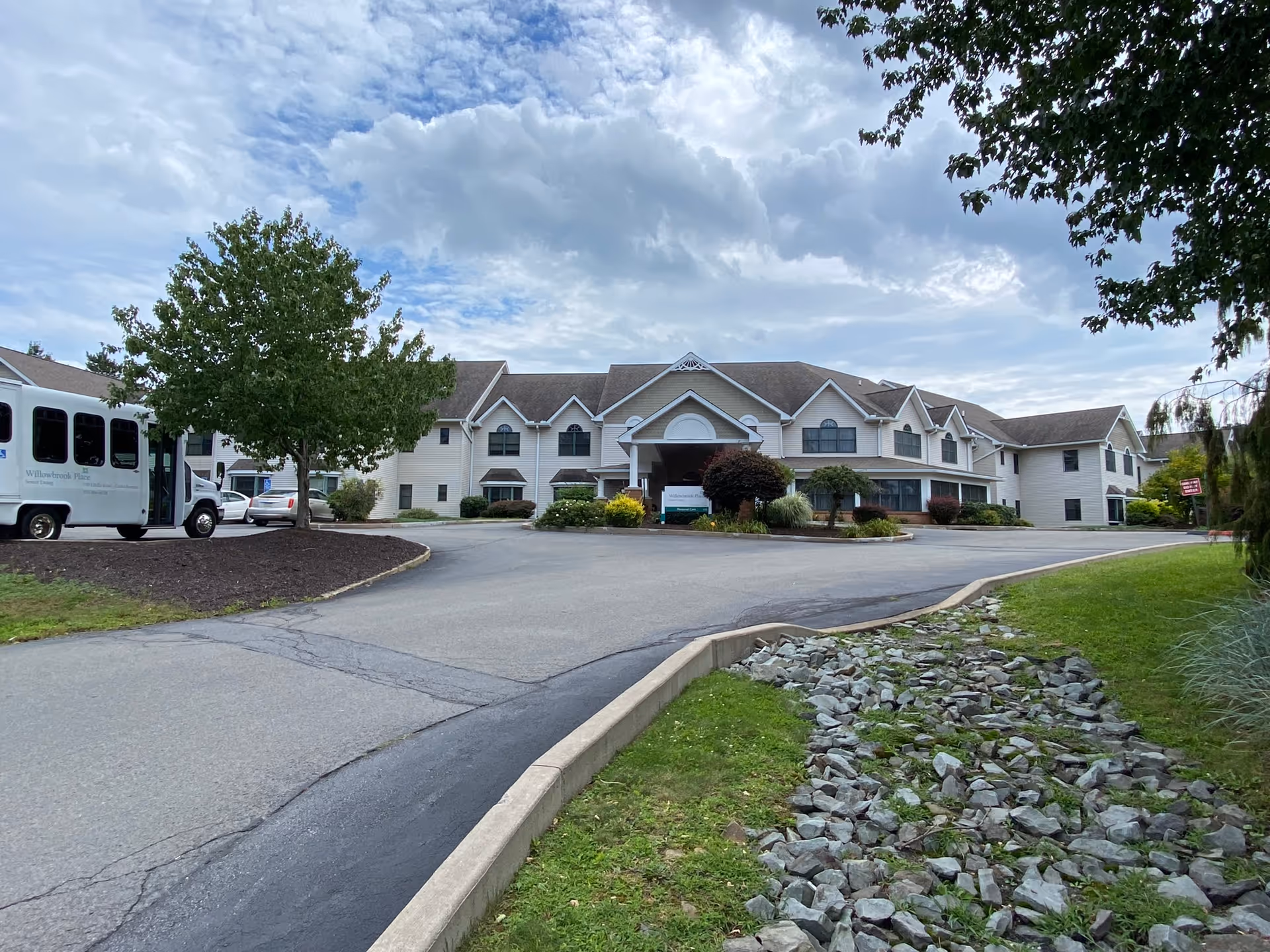 Exterior view of Willowbrook Place, a large two-story senior living facility with a driveway leading to the main entrance. There is a white shuttle bus parked on the left side near a tree, and the sky is partly cloudy.