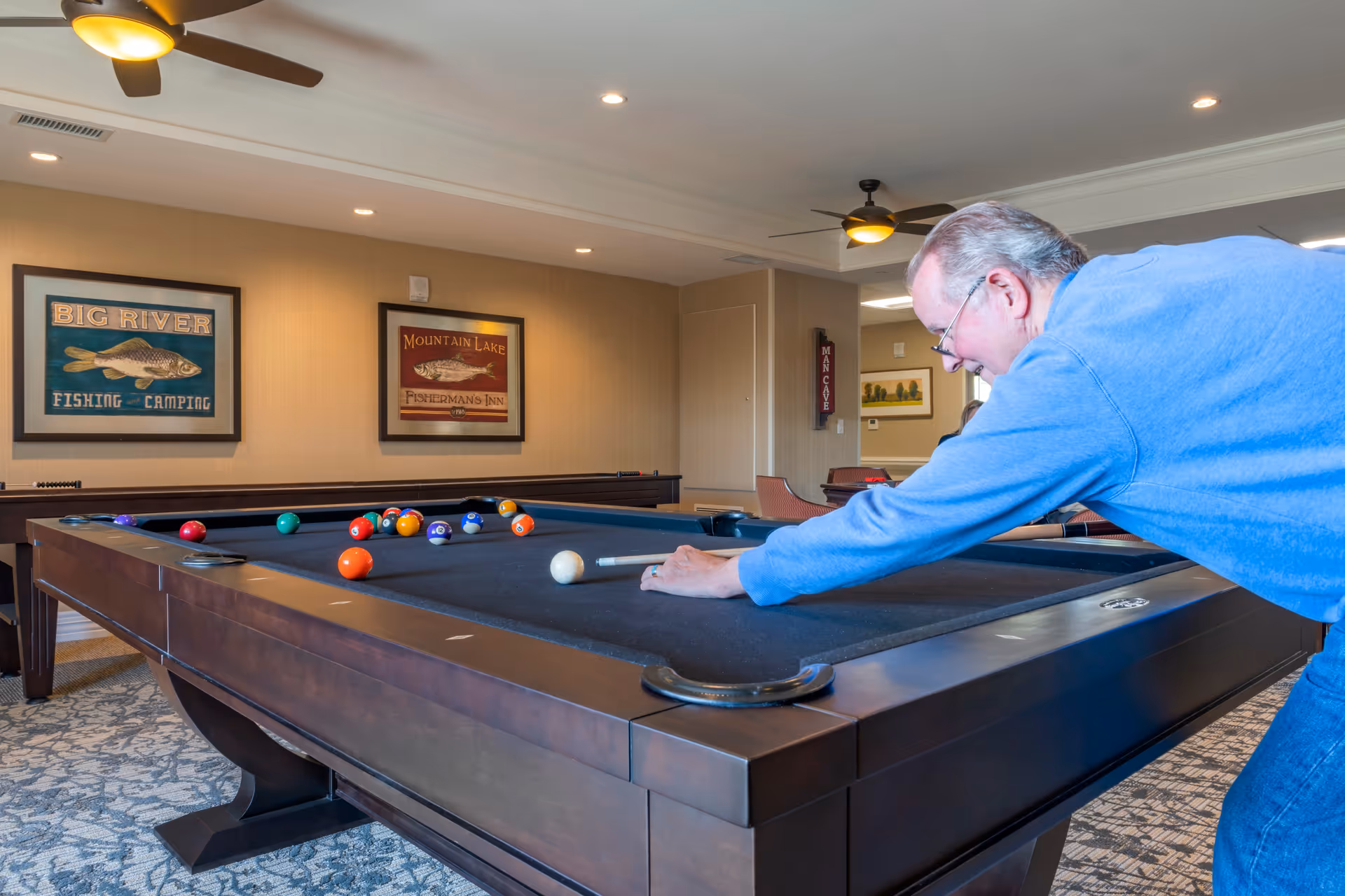 A man lined up to take a shot on a pool table in a well-lit recreation room with ceiling fans and framed fishing artwork.