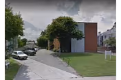 Street view of a brick residential building with a grassy lawn, trees, and parked cars along the driveway.