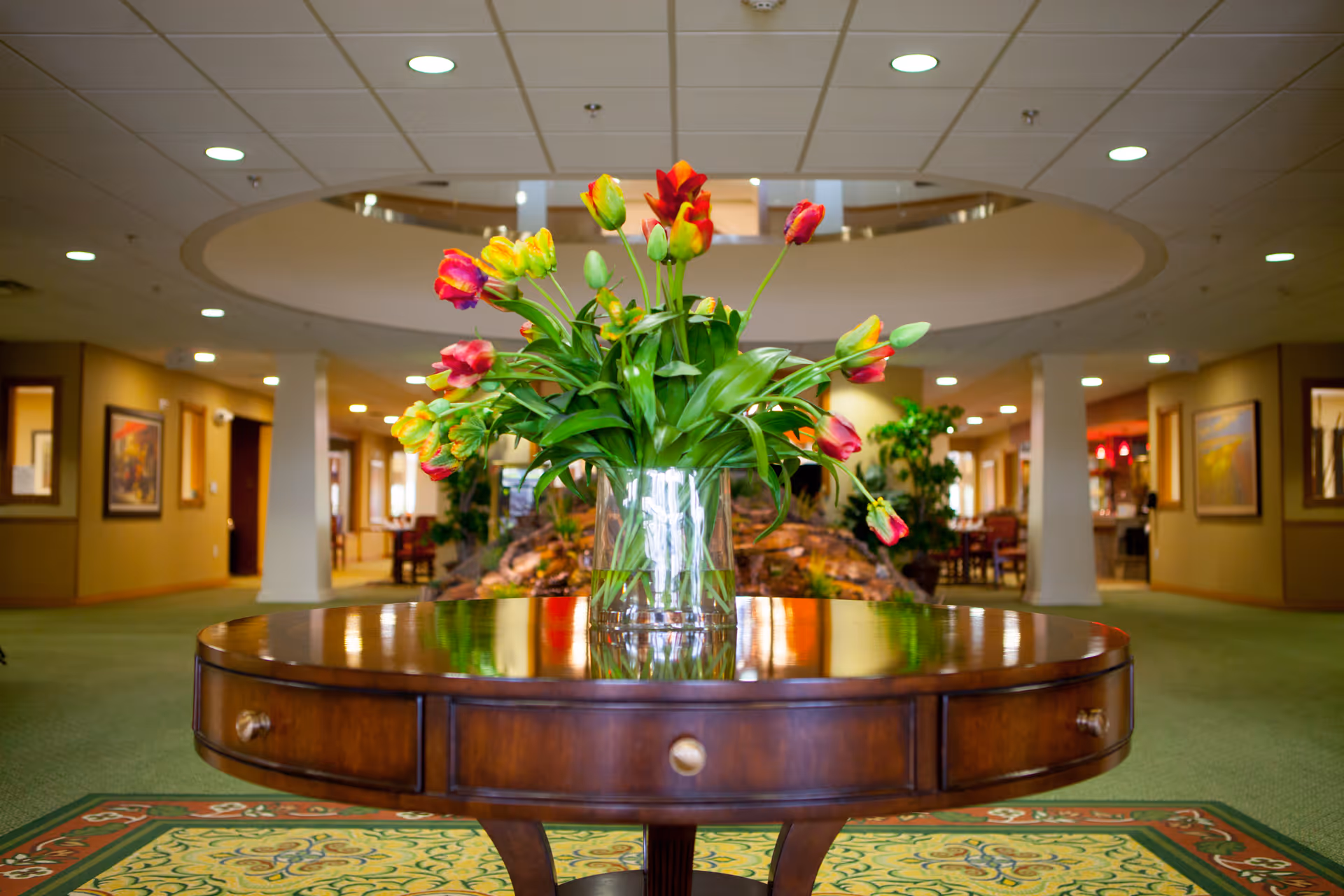 A glass vase of colorful tulips sits on a round wooden table in the lobby of a retirement facility, with seating and decorative lighting in the background.