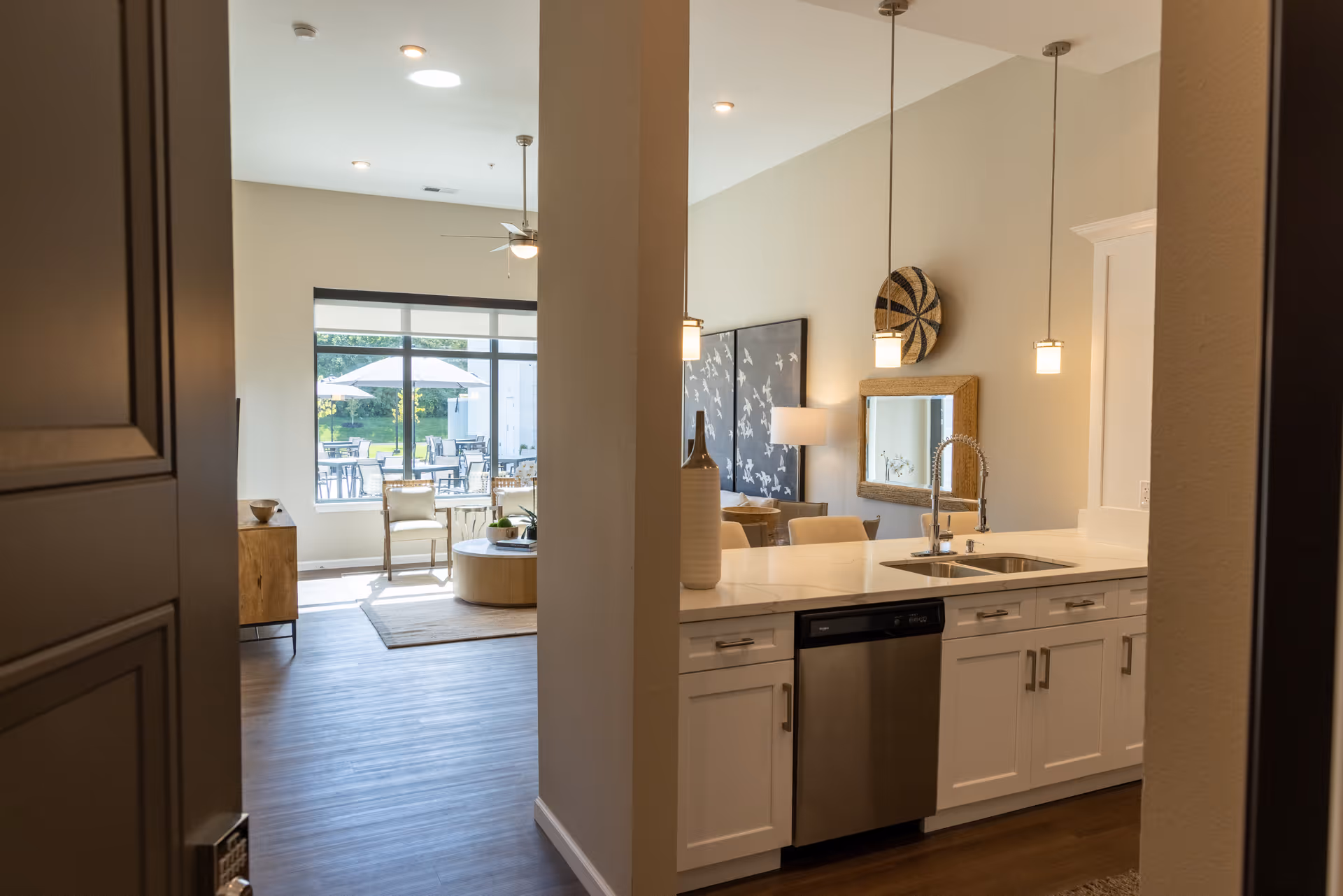 View from an entry into a modern kitchen with a sink island and pendant lights opening to a bright living area and patio seating through large windows.