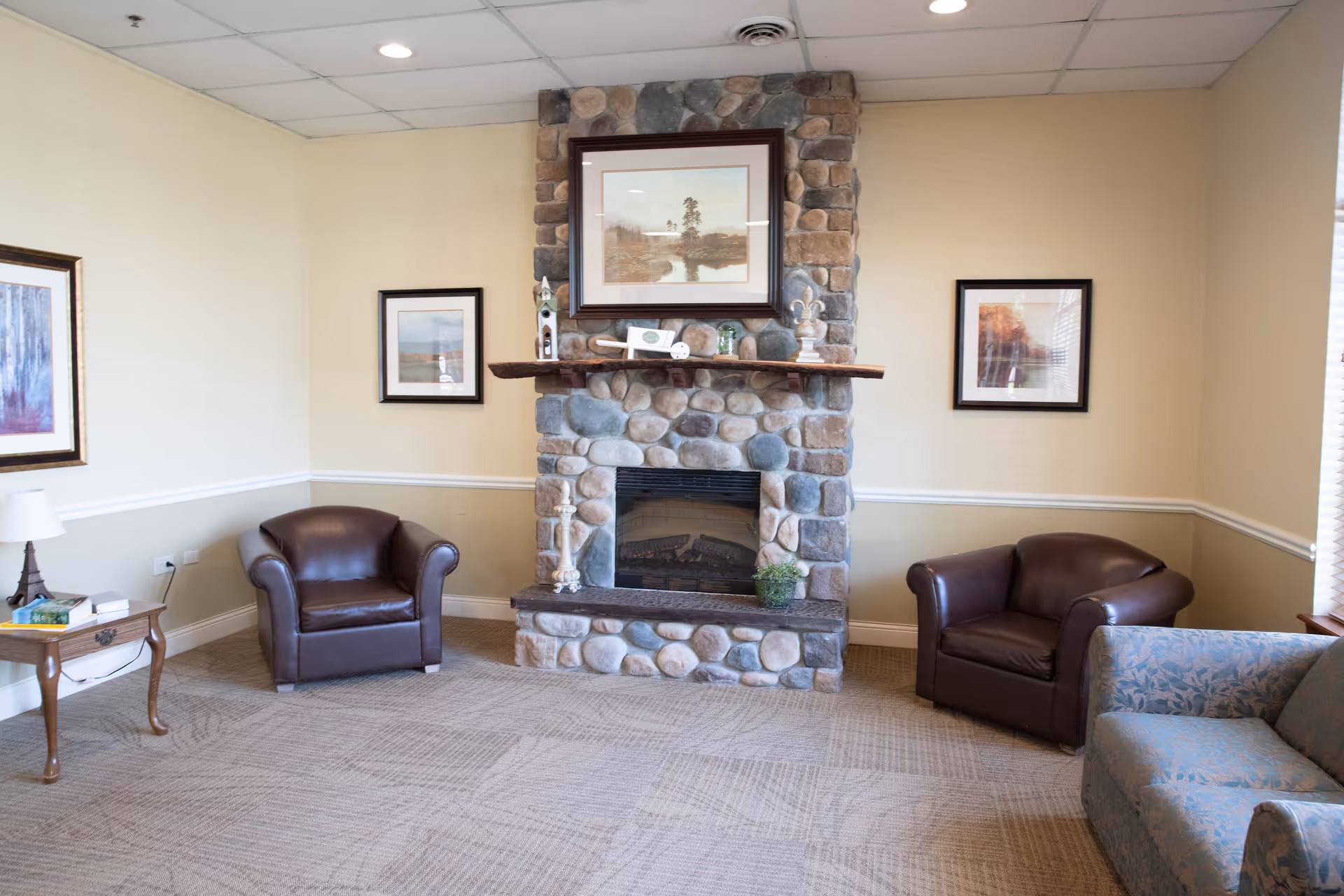 A cozy living room area with a stone fireplace in the center. On either side of the fireplace are two brown leather armchairs, and to the right is a blue patterned sofa. The walls are beige with white trim, and there are three framed pictures hanging on the walls. A small wooden side table with a lamp and some books is on the left side of the image.