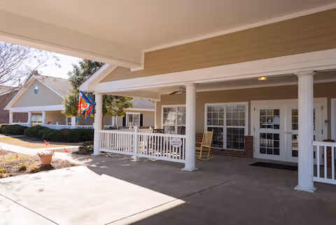 Covered front entrance and porch of a senior living facility with white columns, a rocking chair, and a flag by the walkway.