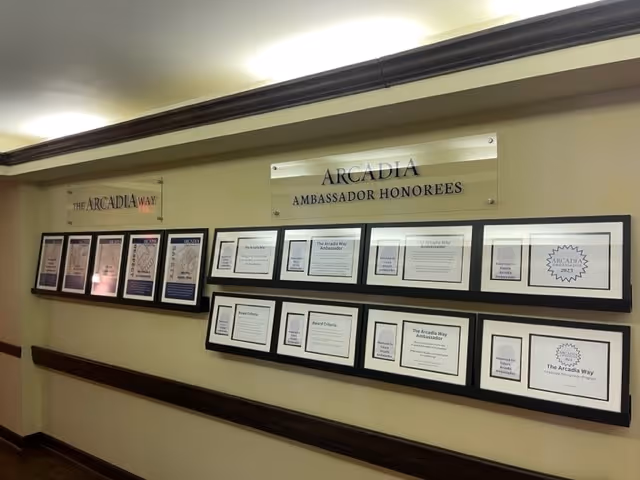 Interior hallway wall with framed certificates and signs reading 'Arcadia Ambassador Honorees' and 'The Arcadia Way'.
