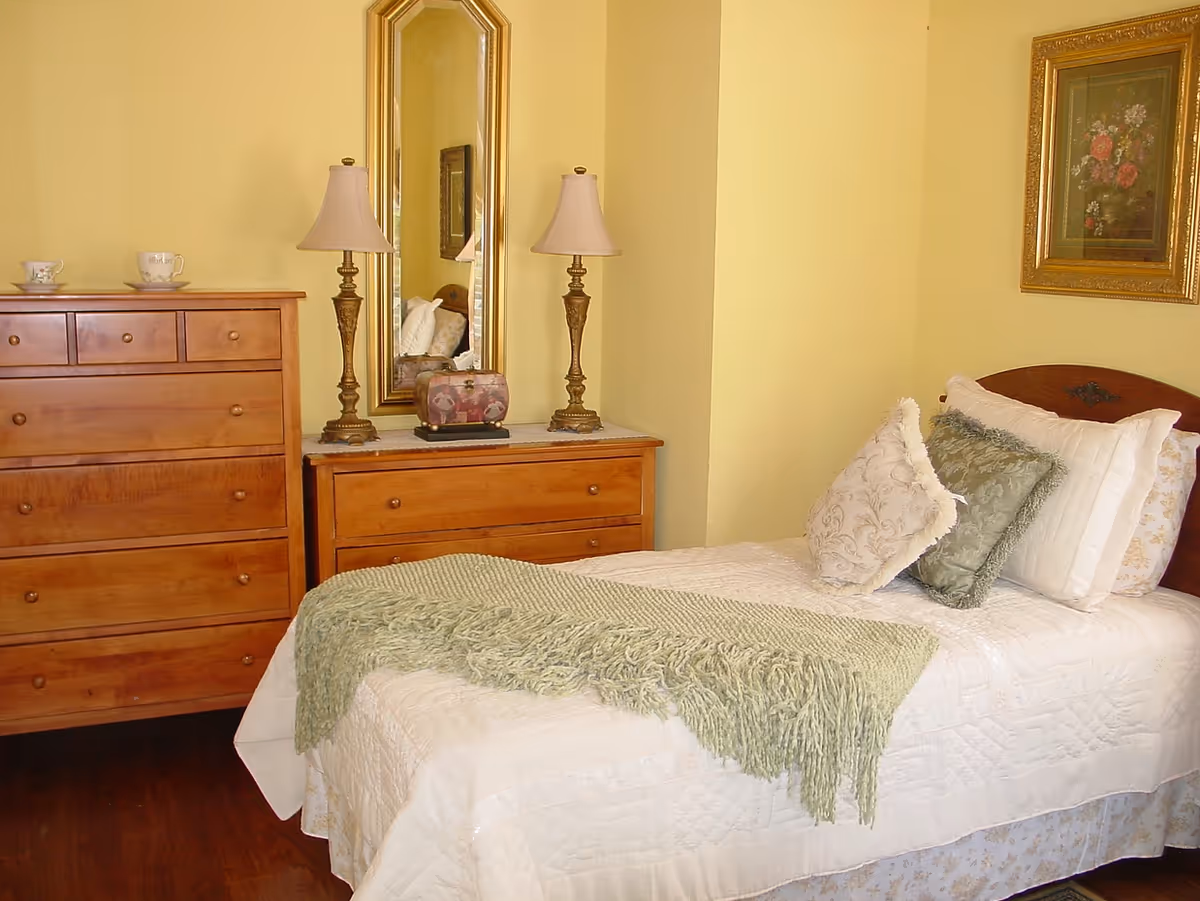 A neatly made single bed with pillows and a green throw in a warmly lit bedroom with wooden dressers, table lamps and a mirror.