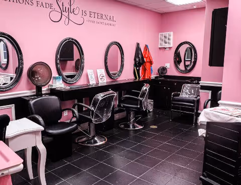 Interior of a beauty salon with pink walls, black tiled floor, and multiple black salon chairs in front of oval mirrors mounted on the wall. Various hair styling tools and products are visible on the counters, along with a quote on the wall that reads 'Fashions fade, style is eternal - Yves Saint Laurent'.