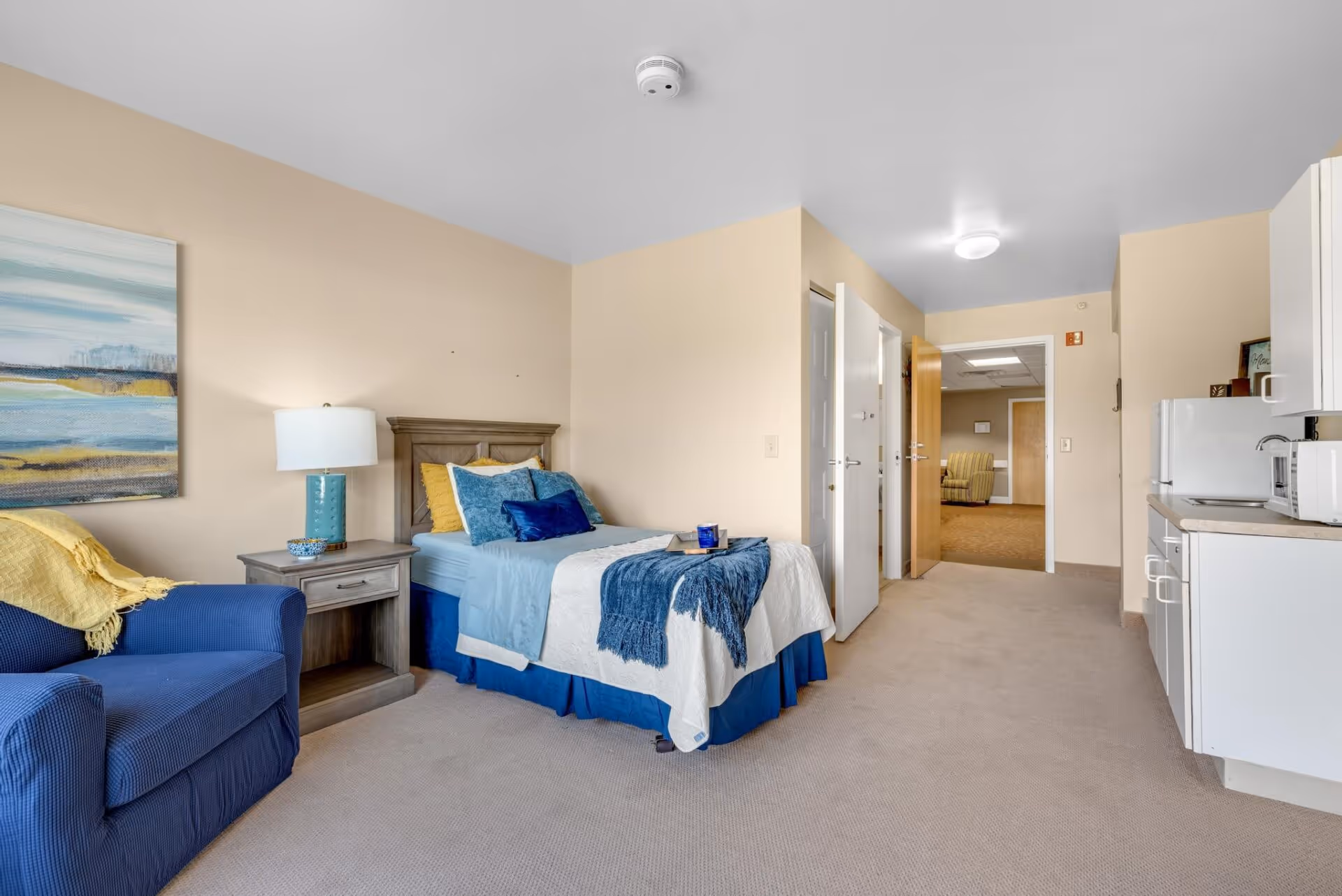 A senior living facility room featuring a single bed with blue and white bedding, a wooden nightstand with a lamp, a blue armchair with a yellow throw blanket, a kitchenette with white cabinets, a microwave, and a refrigerator. The room has beige walls and carpet, and a hallway leading to another room is visible in the background.