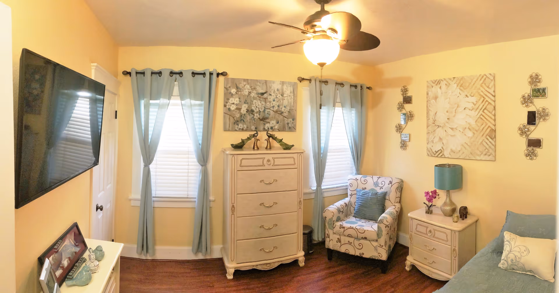Bright bedroom with yellow walls showing a dresser between two curtained windows, an upholstered armchair, nightstand, wall-mounted TV and part of a bed.